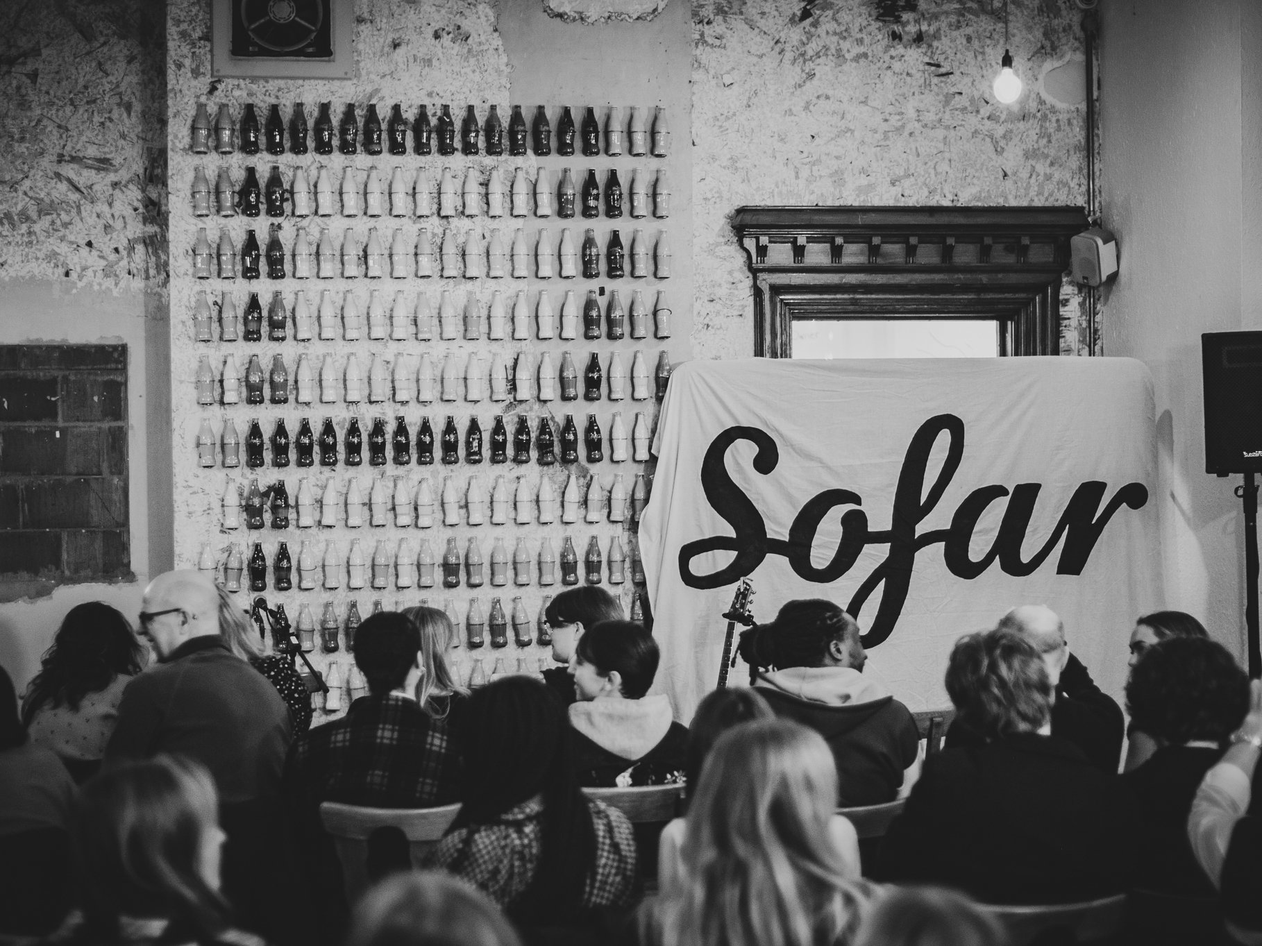 A group of people sitting in front of a stage with a large banner that reads 'Sofar'. The audience is facing the stage, which has a wall decorated with numerous bottles arranged in a grid pattern.