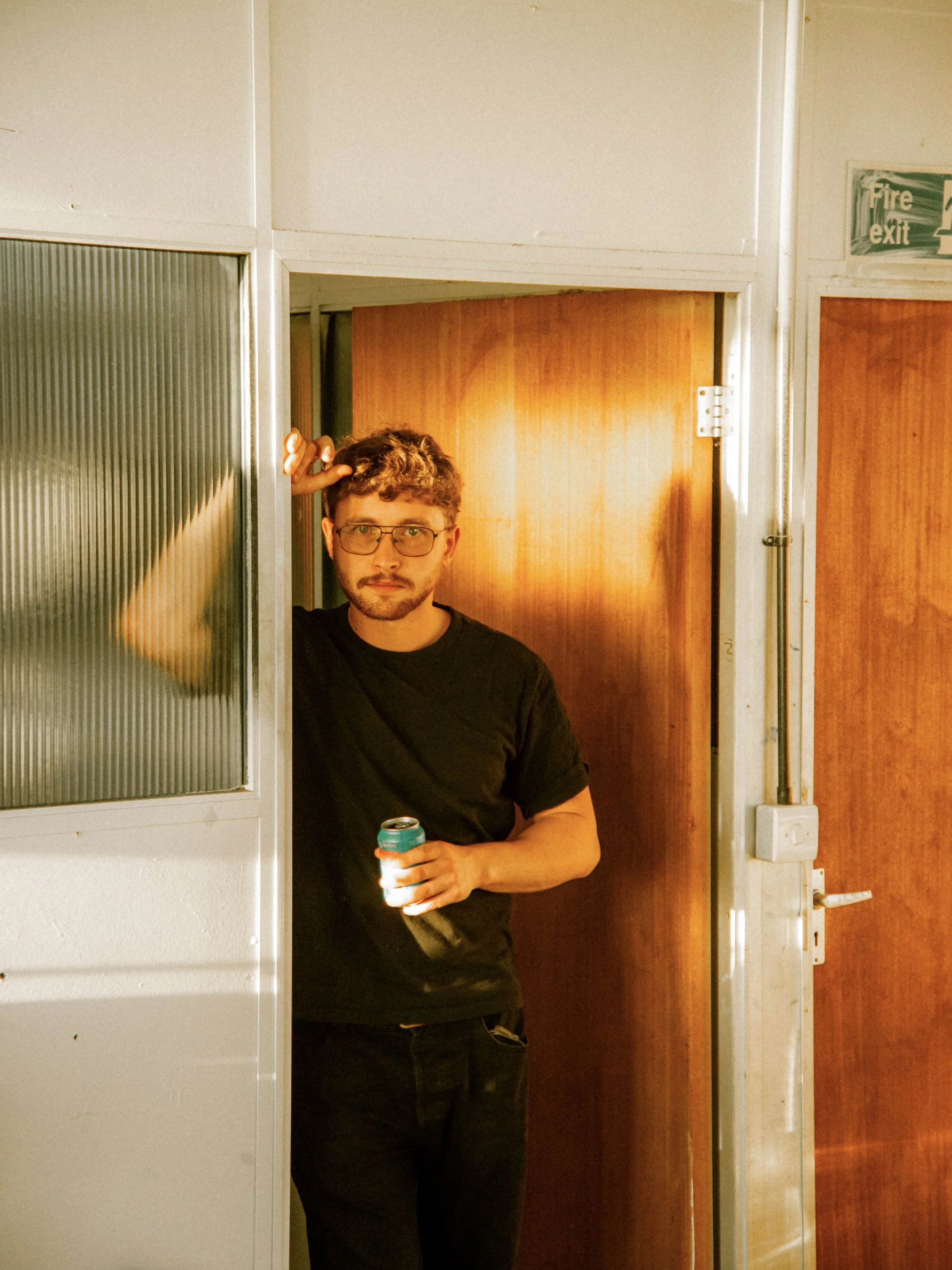 A young man with glasses and curly hair standing in a doorway, holding a drink can, with a neutral expression.