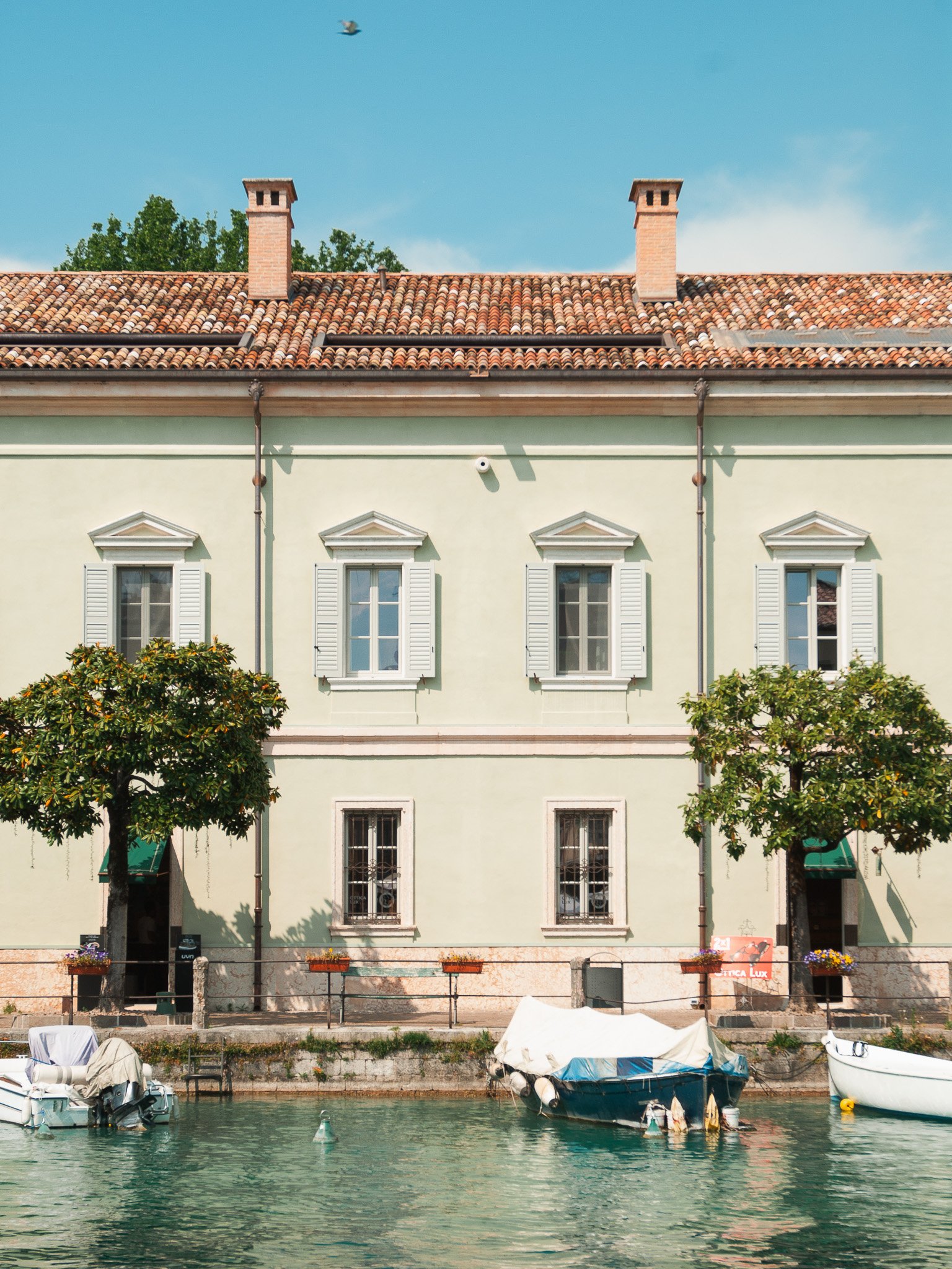 A pastel-colored building along a canal with two trees and boats docked in front, under a clear blue sky.