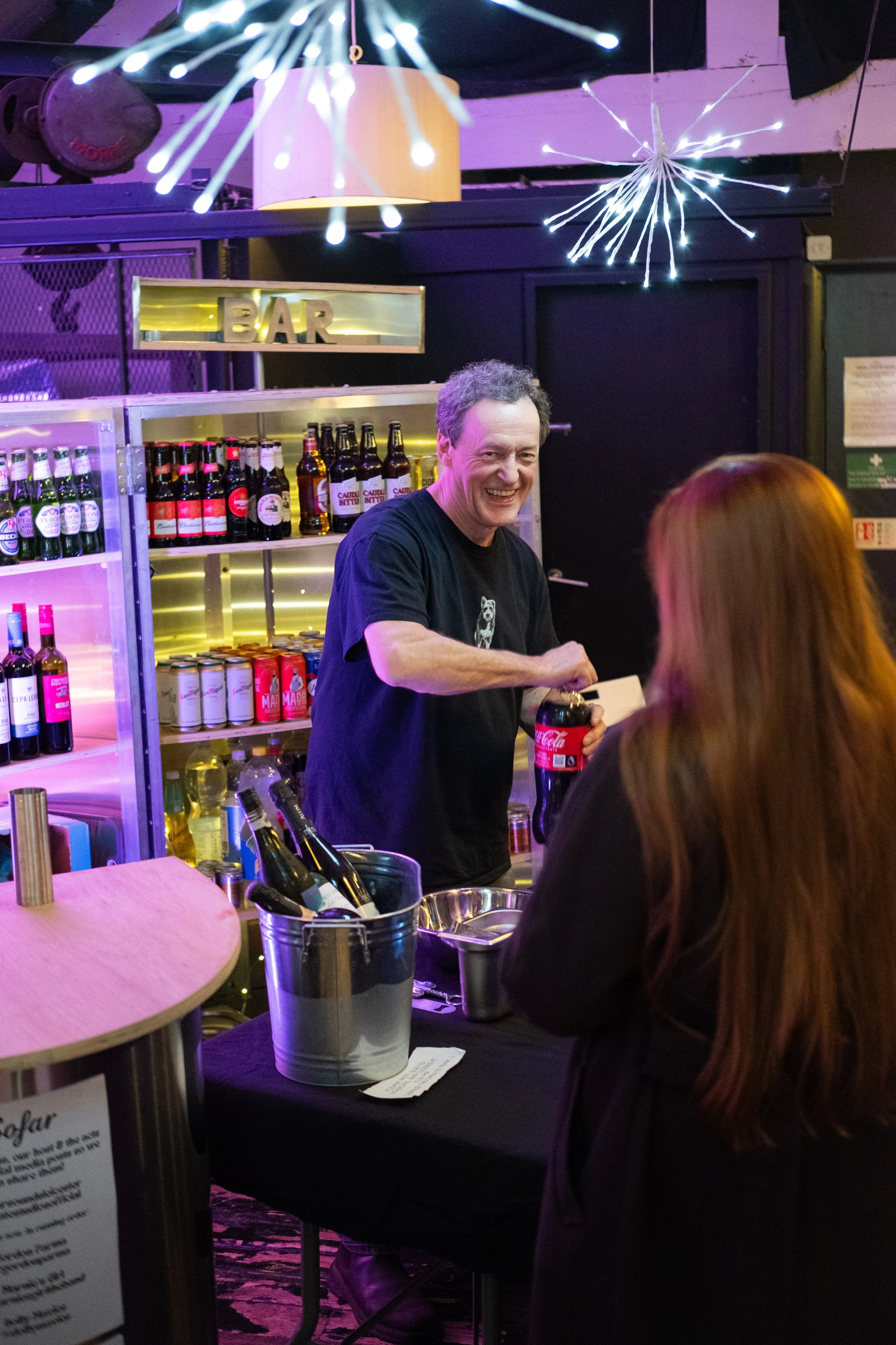 A man with gray hair smiling behind a bar counter, handing a Coca-Cola bottle to a woman with long reddish hair, in a bar with purple and yellow lighting, bottles on shelves, and decorative lighting fixtures hanging from the ceiling.