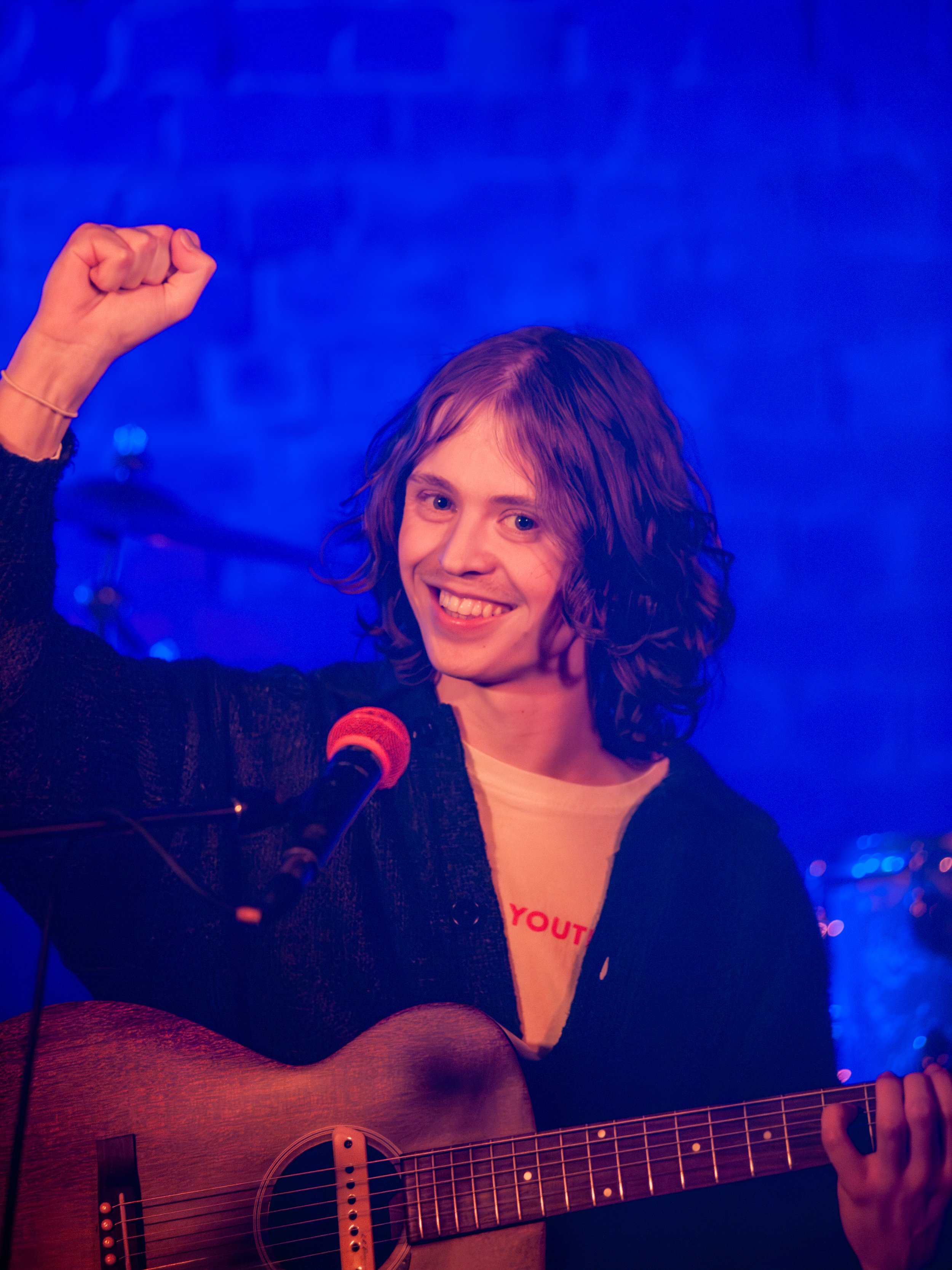 A young woman with shoulder-length curly hair and a bright smile playing an acoustic guitar and raising her right fist in a gesture of victory on stage. She is in front of a microphone and the background features blue lighting and a brick wall.