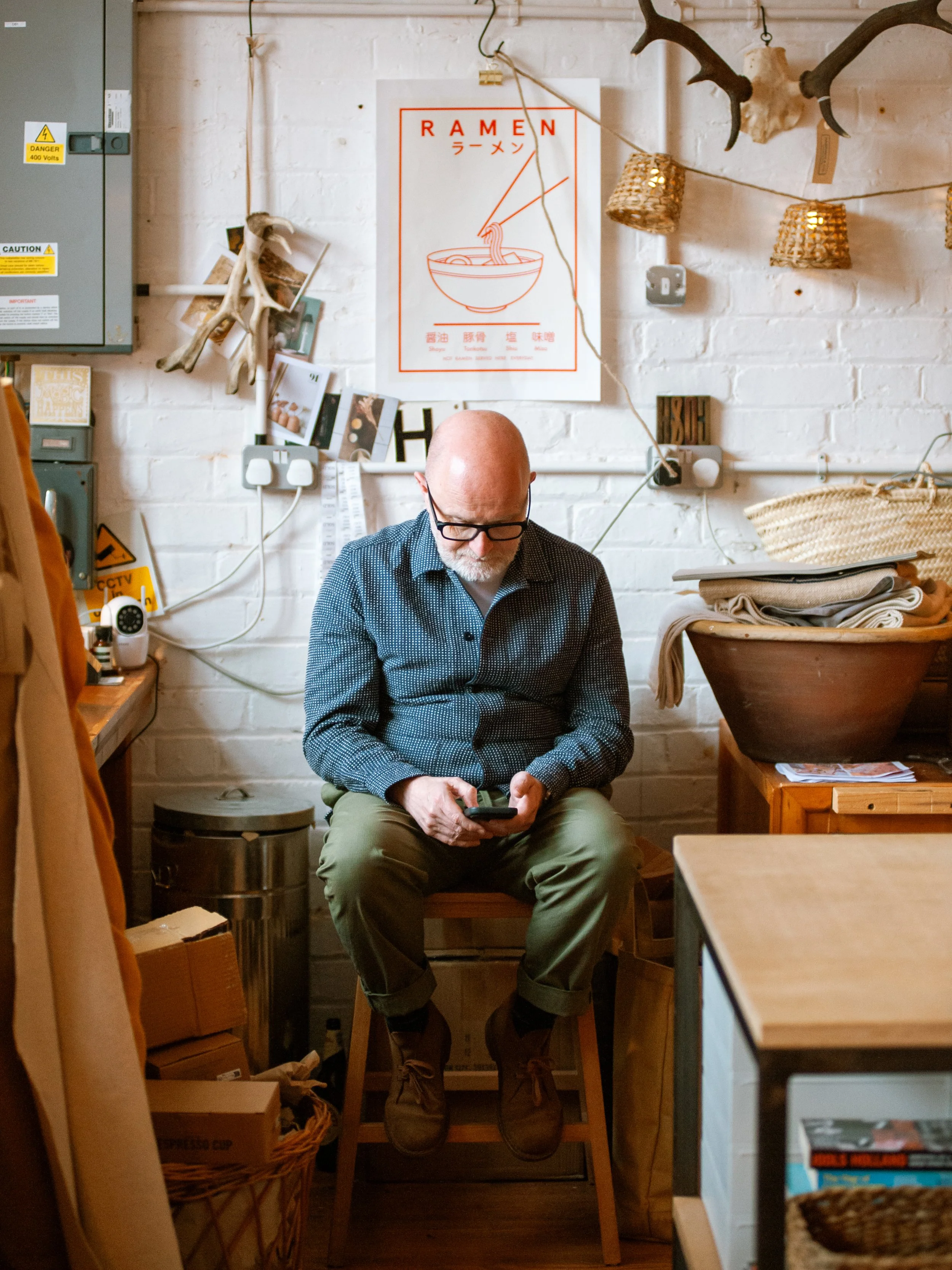 A man with glasses, a beard, and a bald head sitting on a wooden chair in a room, looking at his phone. The room has a white brick wall with a Ramen poster, photos, and decorations. There are various items including a basket, cardboard boxes, and a l