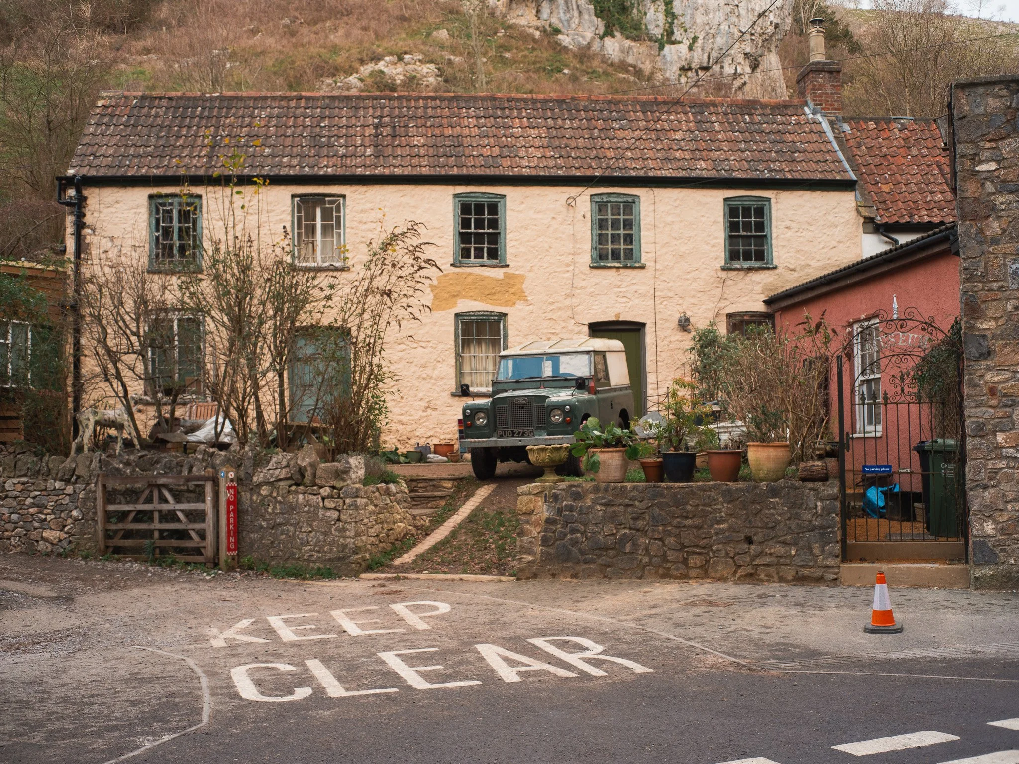A rustic house with a pinkish and beige exterior, multiple windows, and a tiled roof. There are potted plants on the front steps and a vintage truck parked in the yard. A traffic cone and a 'Keep Clear' sign are painted on the street in front of the 