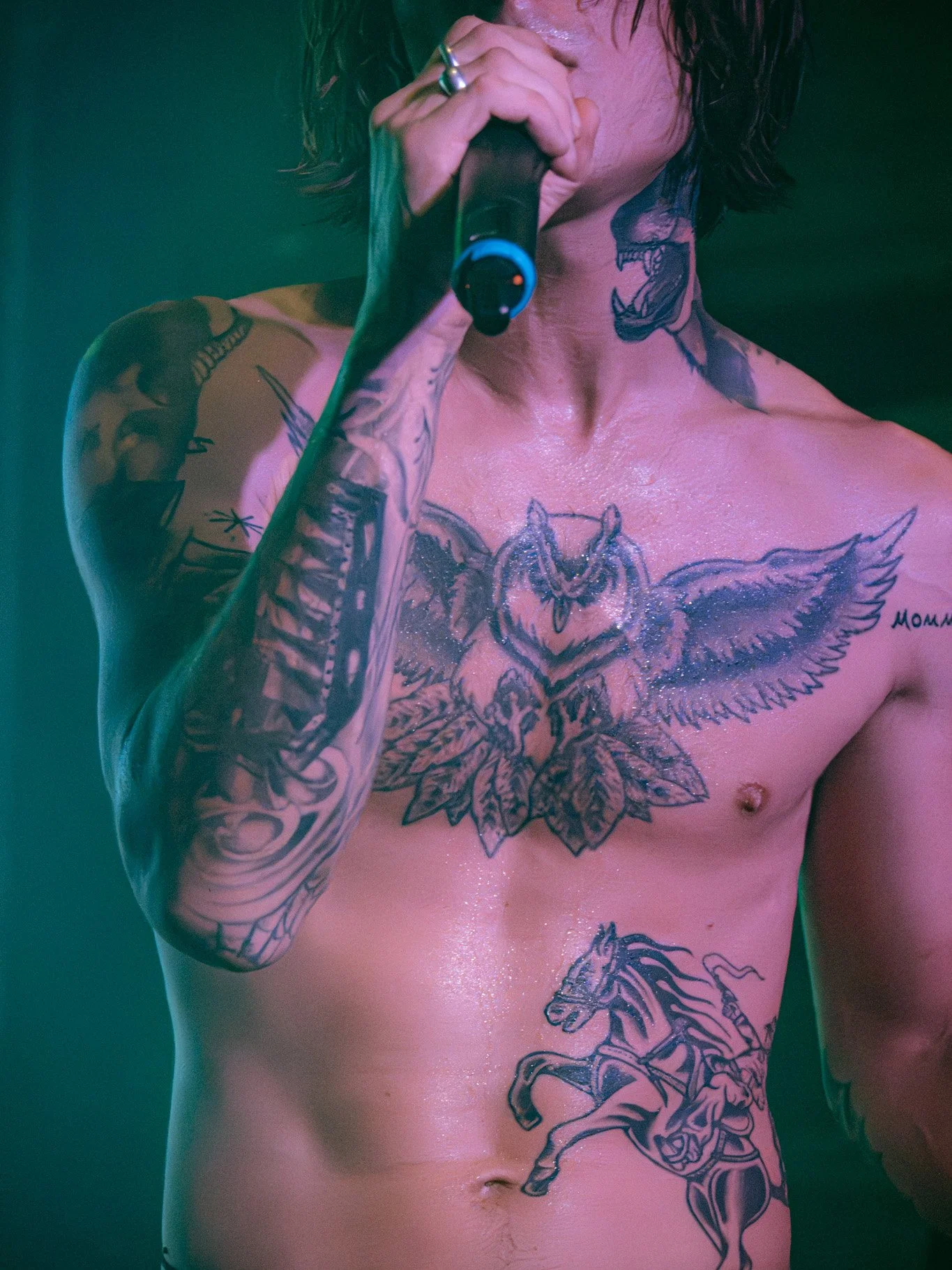 A shirtless man with dark, curly hair holding a microphone. He has multiple tattoos, including a large owl with spread wings on his chest, a horse on his stomach, and various tattoos on his arms and neck.