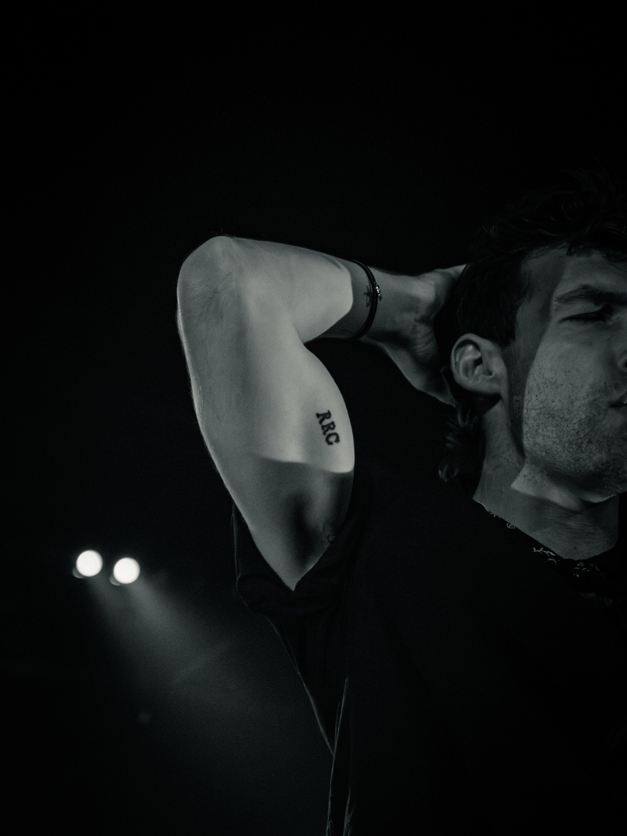 Black and white photo of a man with a tattoo on his arm, holding his head with one hand, on a dark background with spotlights in the distance.