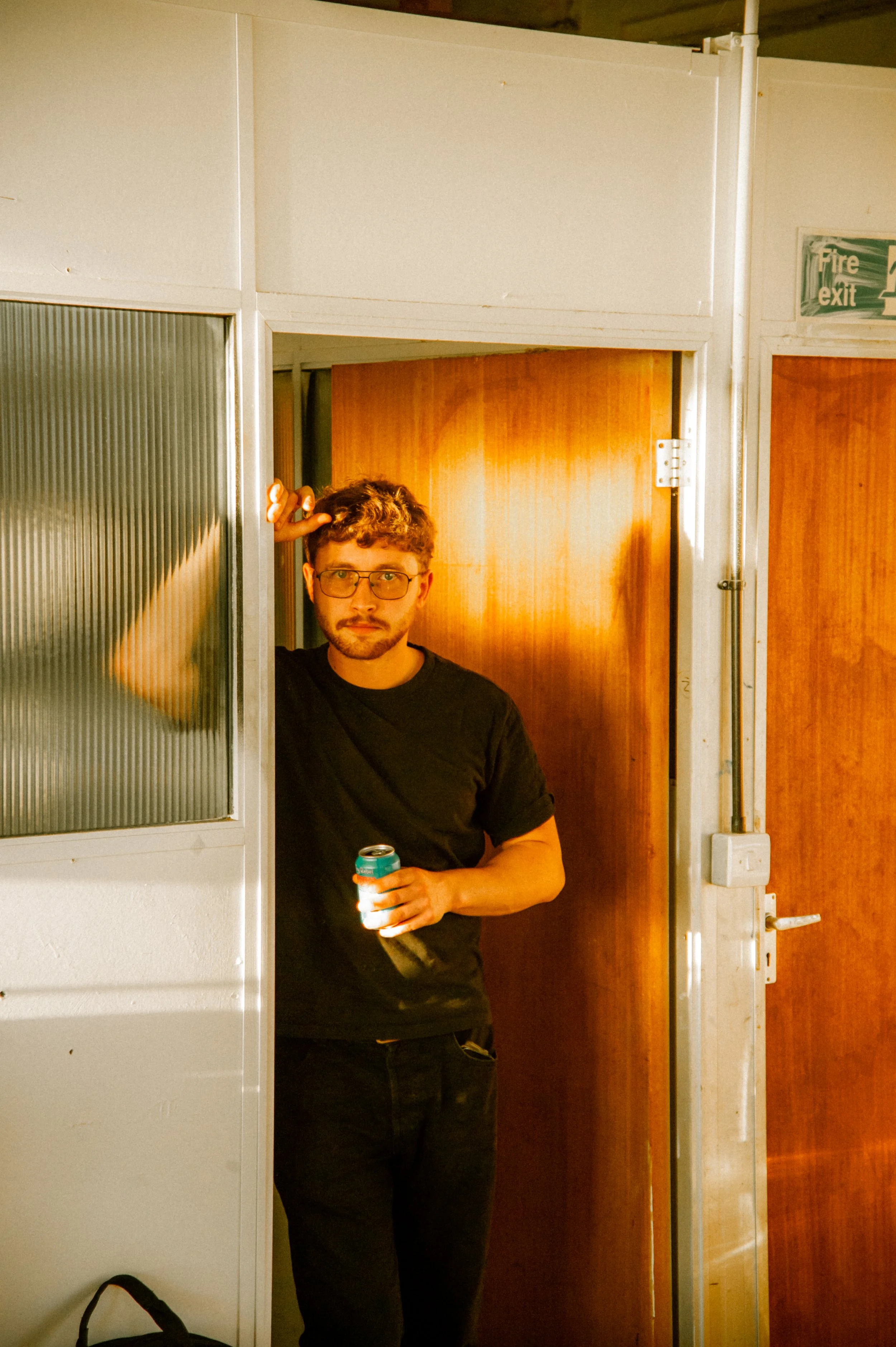 A young man with glasses and a beard, wearing a black t-shirt, stands in a doorway holding a can of drink in his right hand and scratching his head with his left hand. Behind him are wooden doors and an emergency exit sign.
