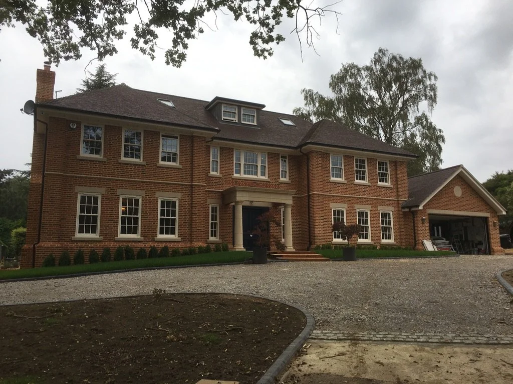 A large brick house with white-framed windows, a garage on the right, and a gravel driveway in the front, surrounded by trees and greenery.