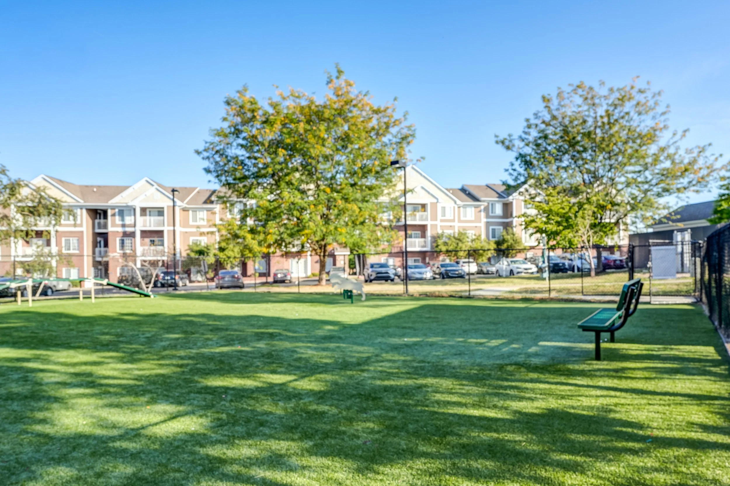 A park with green grass, trees, benches, and a fence with apartment buildings in the background under a clear blue sky.