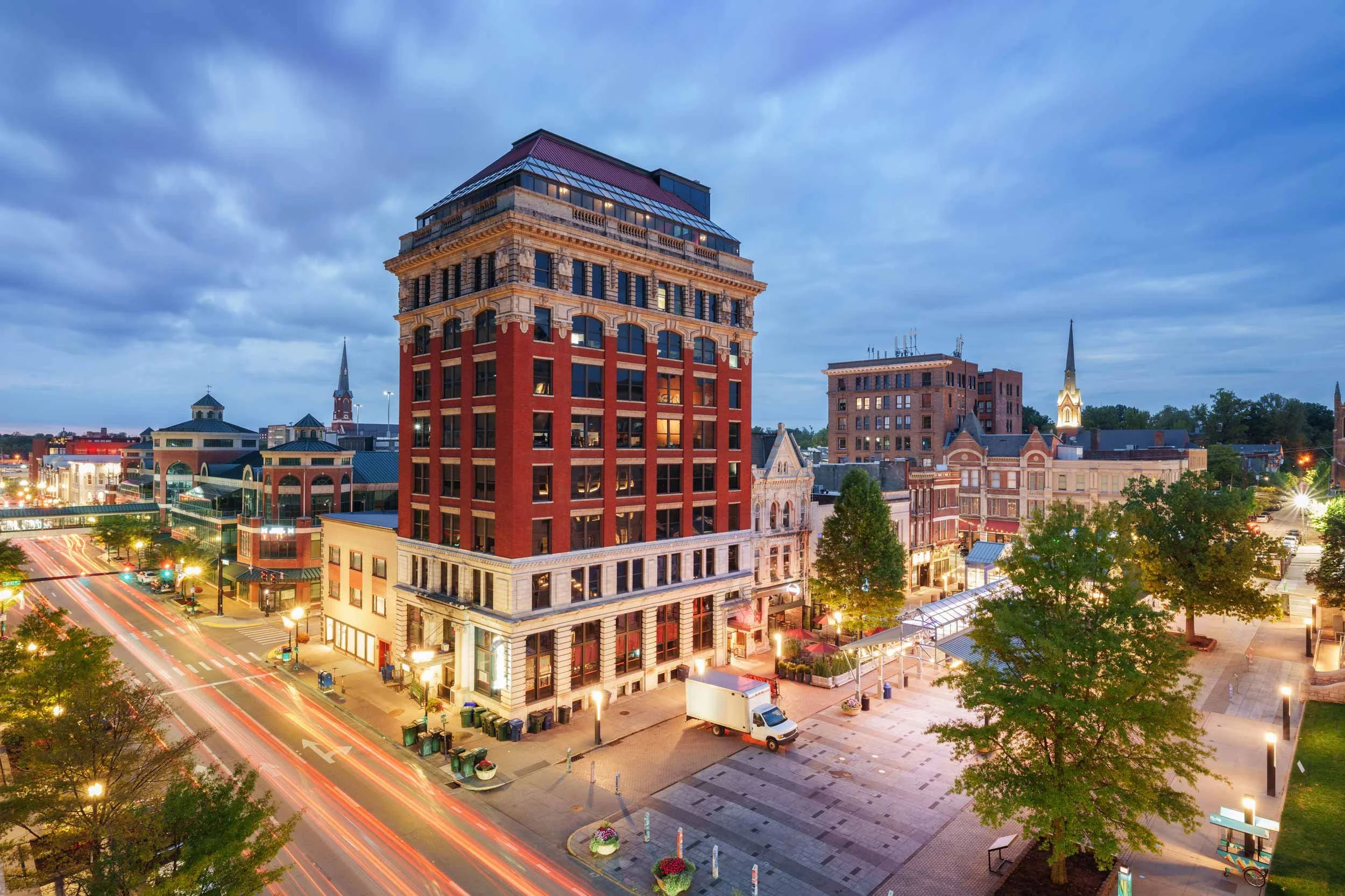 Cityscape during dusk featuring a tall historic brick building with a mansard roof, other brick and stone buildings, church steeples, and illuminated streets with light trails from moving vehicles.