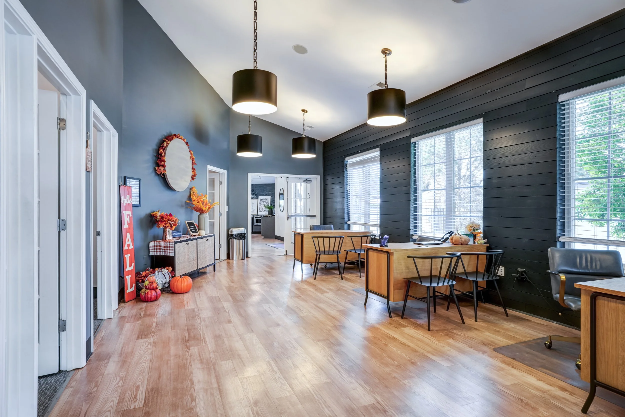 Interior of a cozy cafe or coffee shop with large windows, wooden floors, black and gray walls, and black pendant lights hanging from the ceiling. Decor includes autumn-themed pumpkins and flowers near the doorway, and tables with chairs for seating.