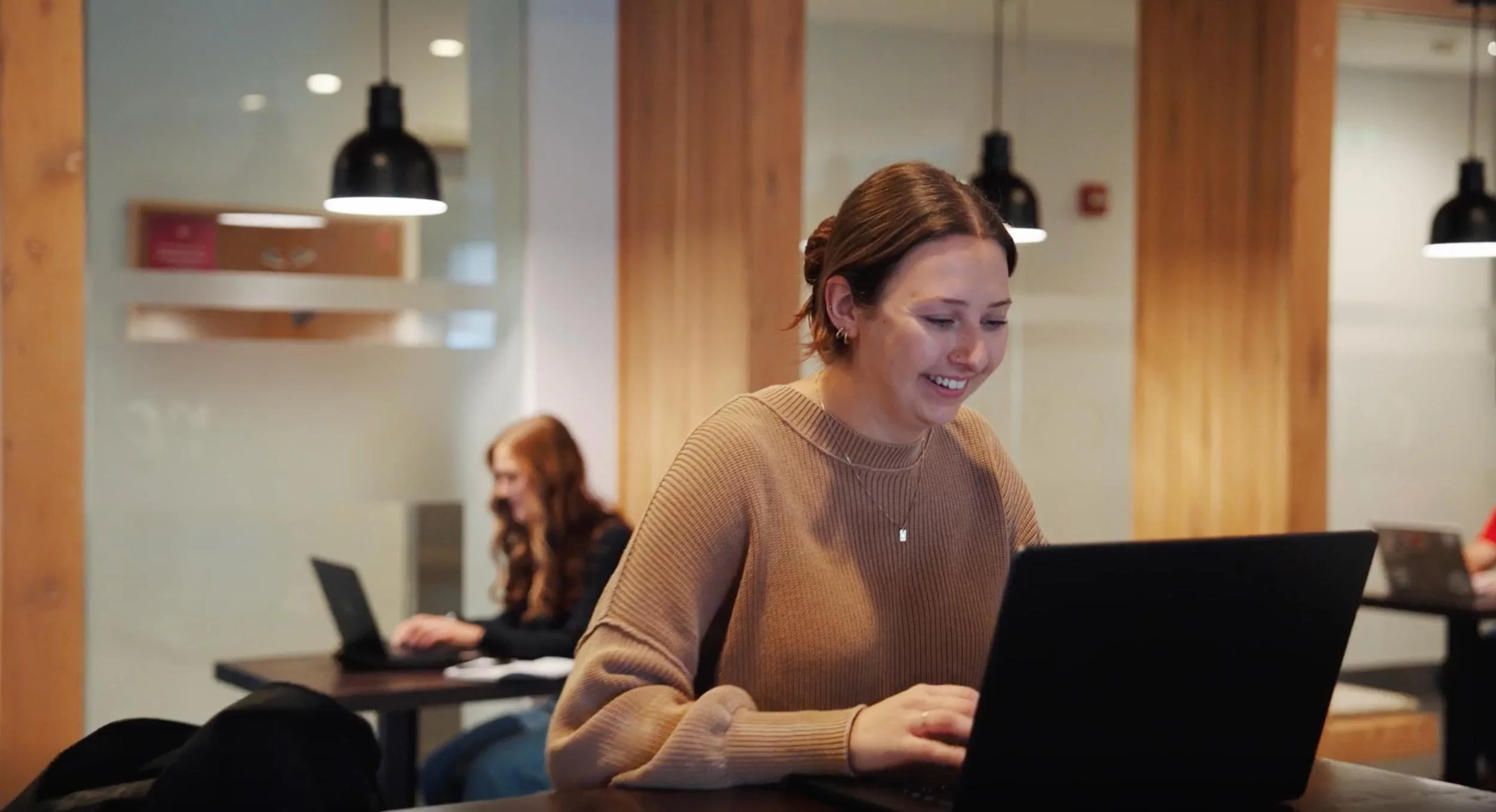A young woman with short brown hair and earrings smiling while working on a black laptop in a coffee shop or coworking space. In the background, another woman with long red hair also working on a laptop at a different table.