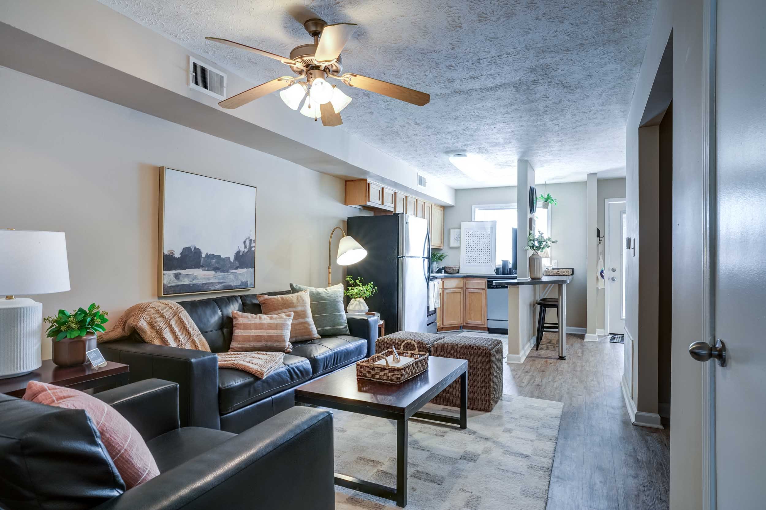 Living room with a black leather sofa, wooden coffee table, and beige pillows. The room has a white and beige color scheme with artwork on the wall, a table lamp, and it opens into a kitchen with wooden cabinets, a refrigerator, and a window letting in natural light.