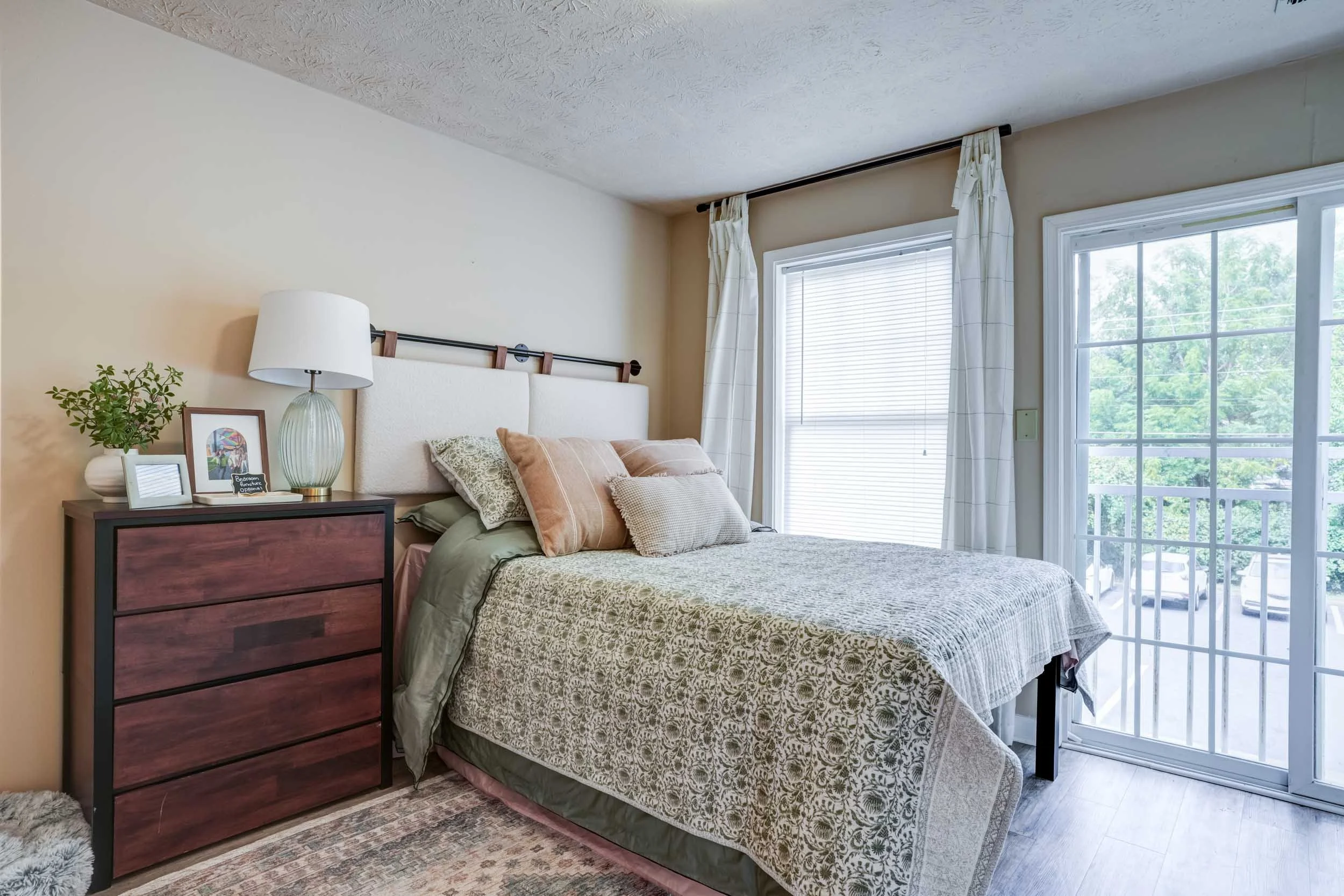 A bedroom with a bed, nightstand, and large windows with white curtains, overlooking trees outside.