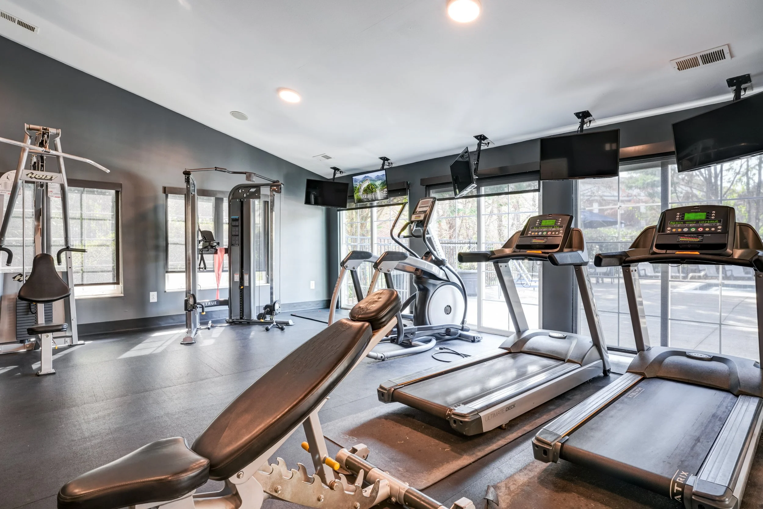 A gym with treadmills, exercise bikes, weightlifting equipment, multiple TV screens, and large windows letting in natural light.