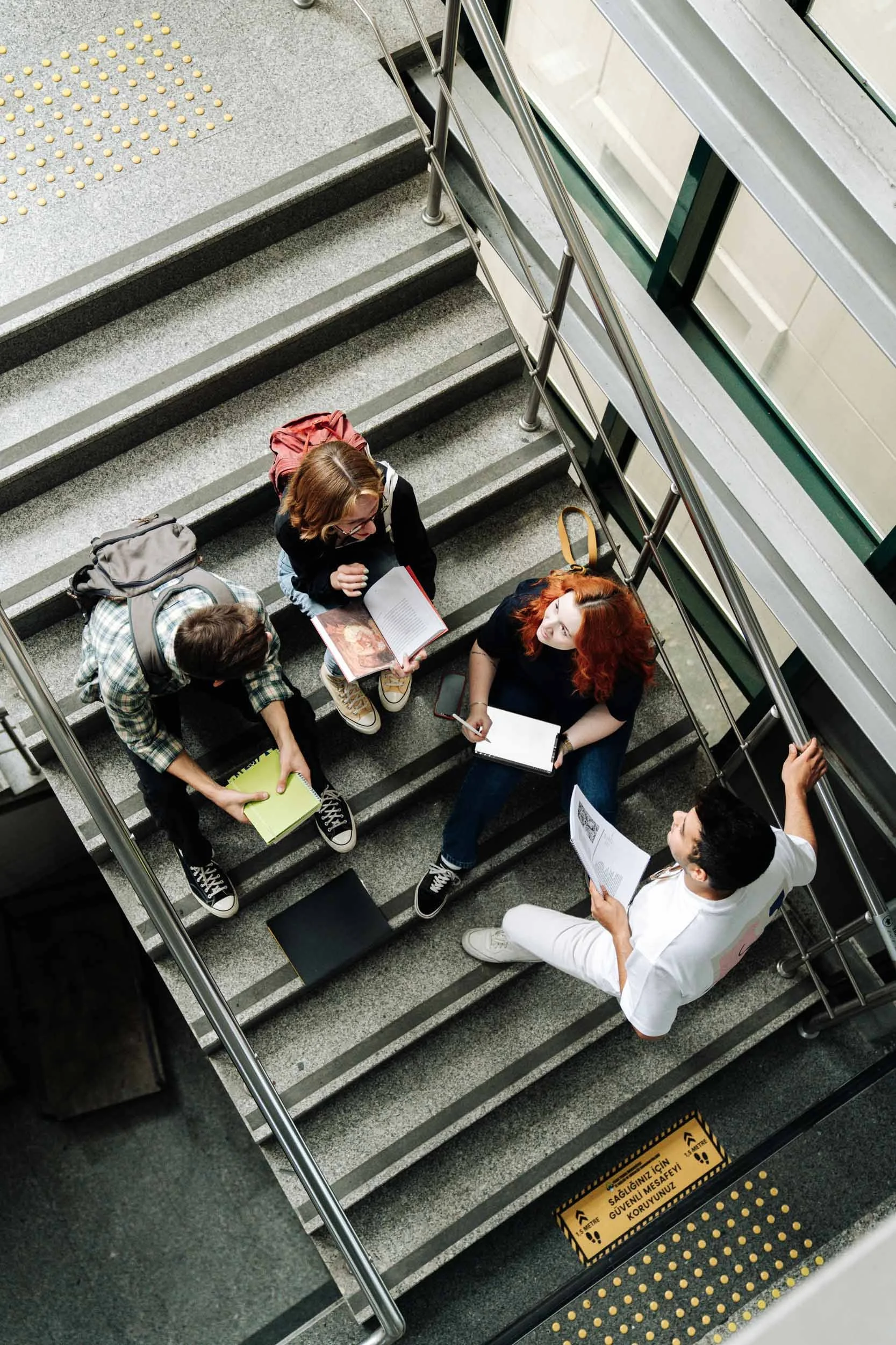 Group of people sitting on stairs inside a building. One person appears to be talking to a group, while others are listening and reading. There are backpacks and notebooks with them. A yellow caution sign is on the stairs, warning about safety.