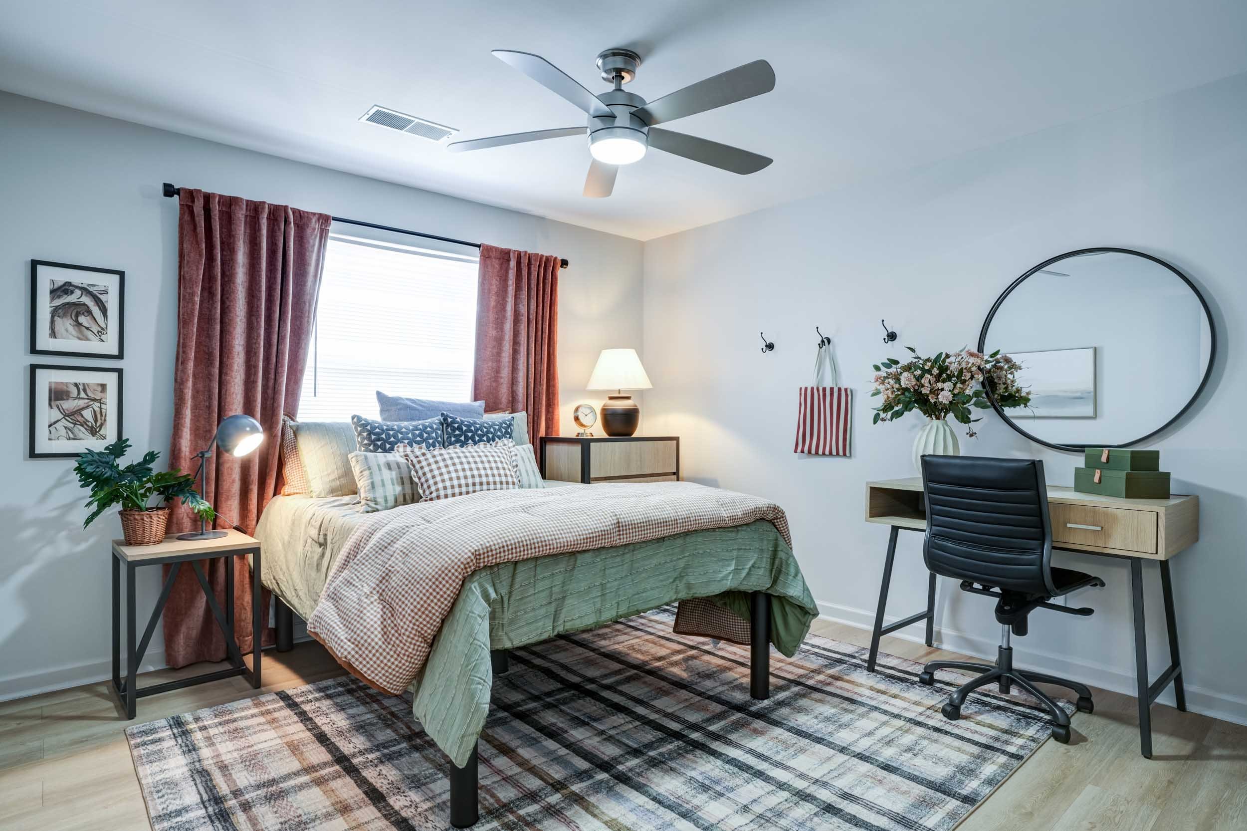 A bedroom with a bed, side table, lamp, window with pink curtains, a desk with a chair, a circular mirror, and a ceiling fan.