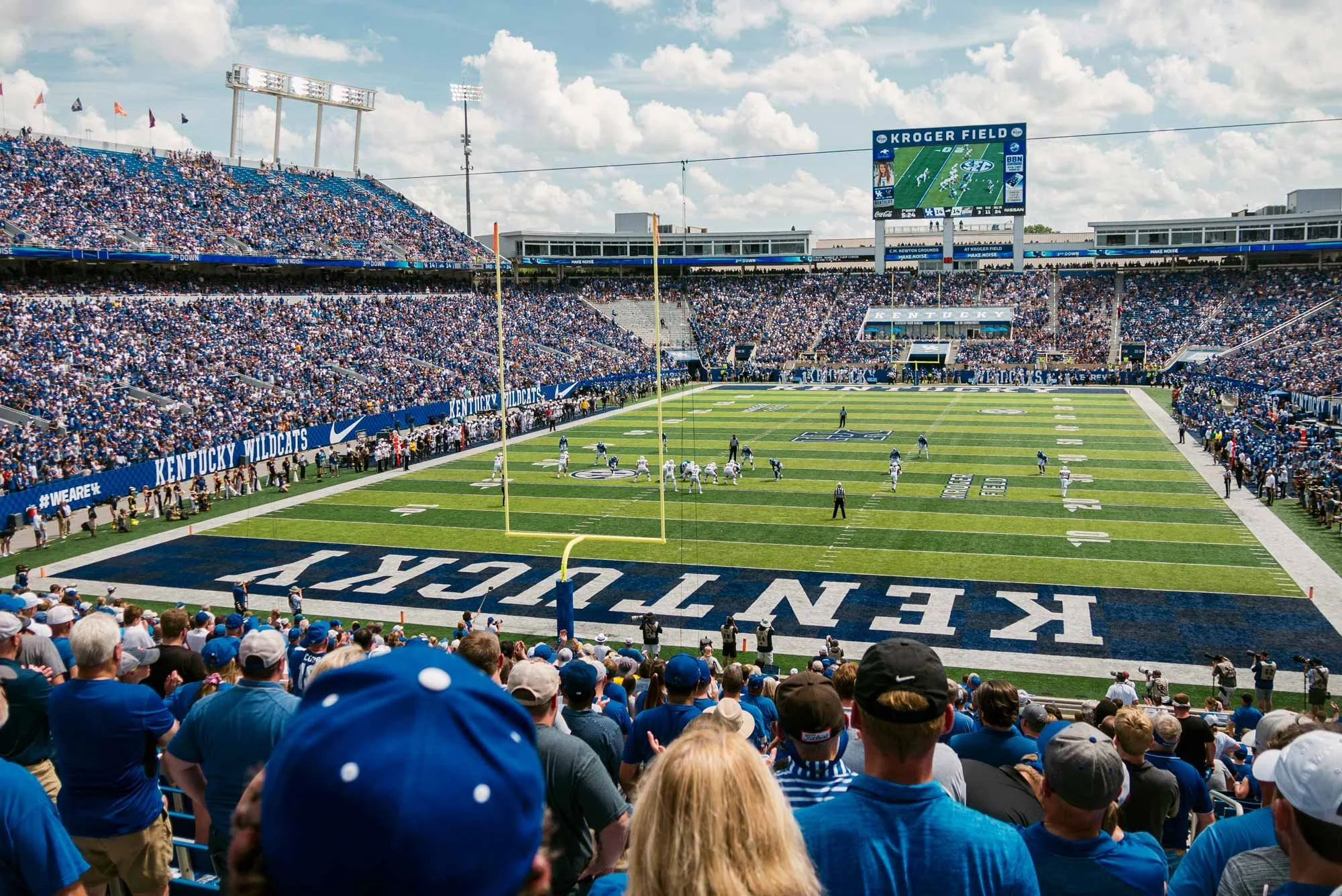 Crowd of spectators watching an American football game at Kroger Field, Kentucky Wildcats team on the field, stadium filled with fans dressed predominantly in blue and white, large scoreboard displays game information, sunny weather with scattered clouds.