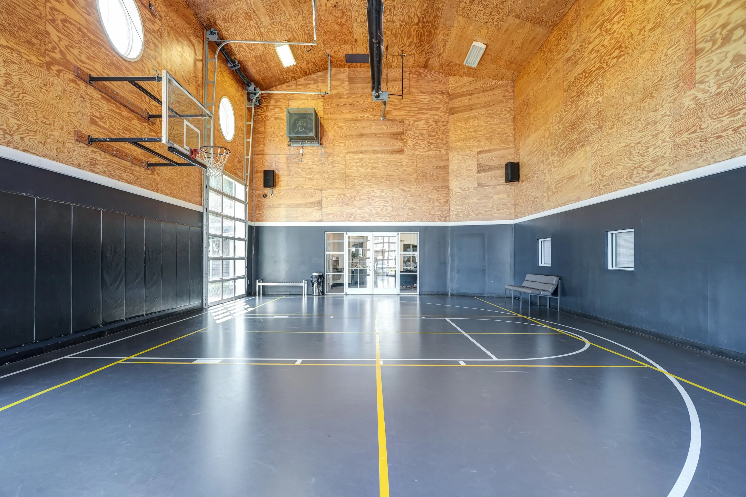 Indoor gymnasium with a wooden back wall, black padded walls, and basketball court markings, including a hoop, some basketball equipment, a bench, and windows.