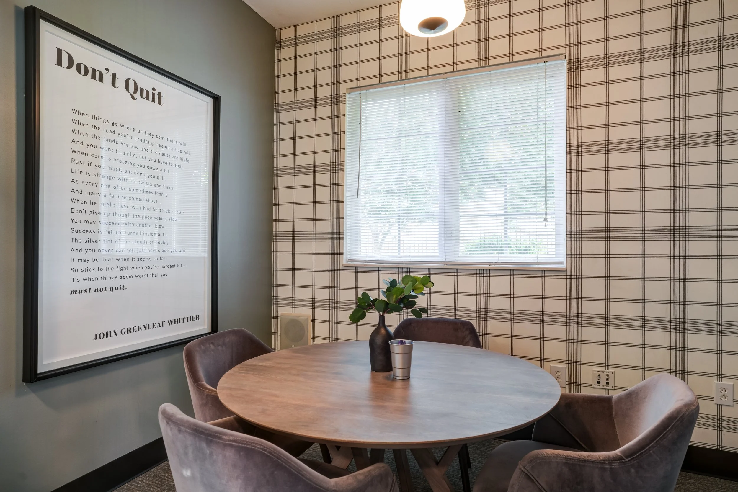 A cozy dining area with a round wooden table surrounded by velvet chairs. On the table, there is a metallic cup and a dark-colored vase with green leaves. The wall features a large framed motivational quote titled 'Don't Quit' and there is a window w