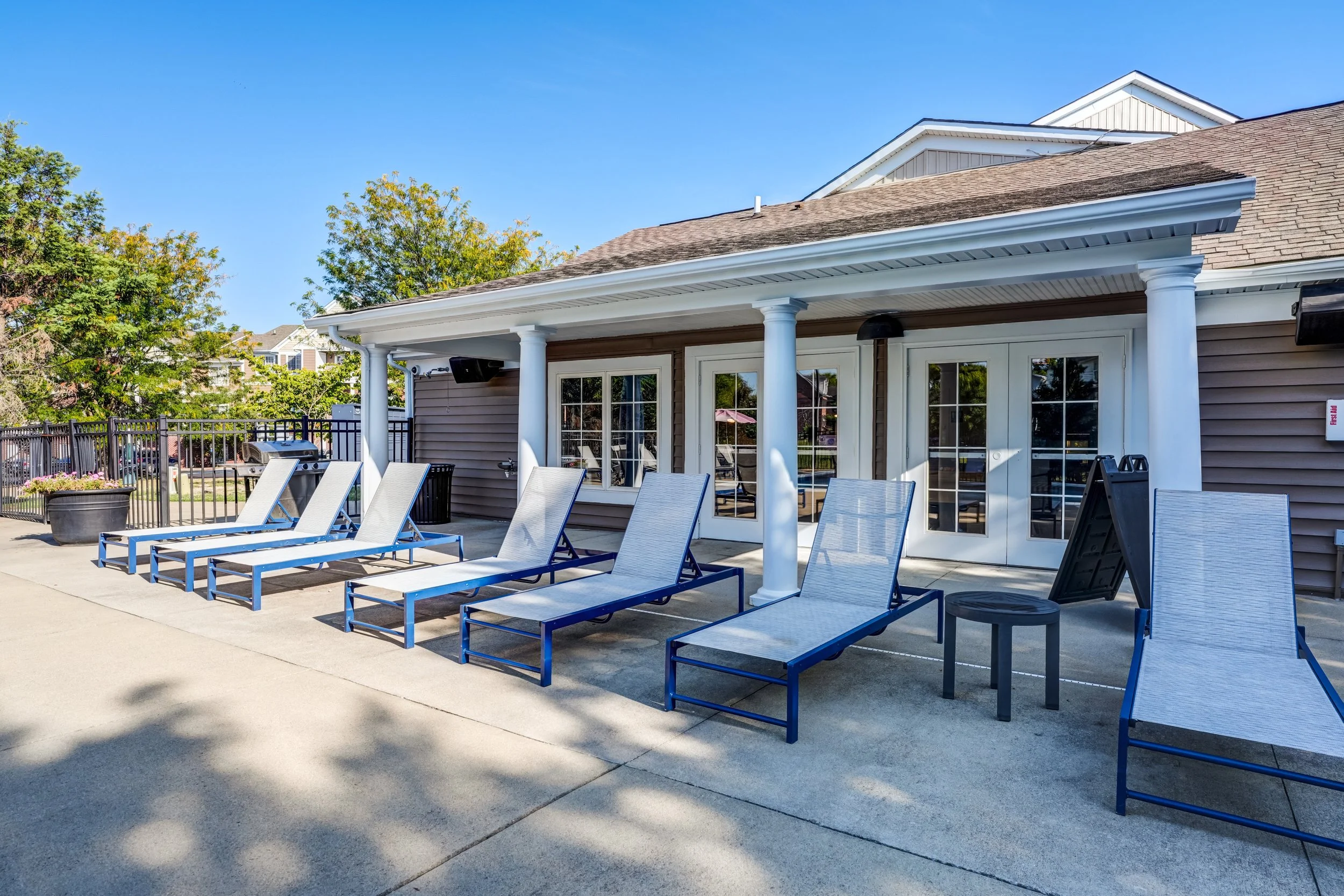 Patio area with multiple white lounge chairs lined up on a concrete surface, adjacent to a house with large glass doors, and a black barbecue grill, under a covered porch with white columns.