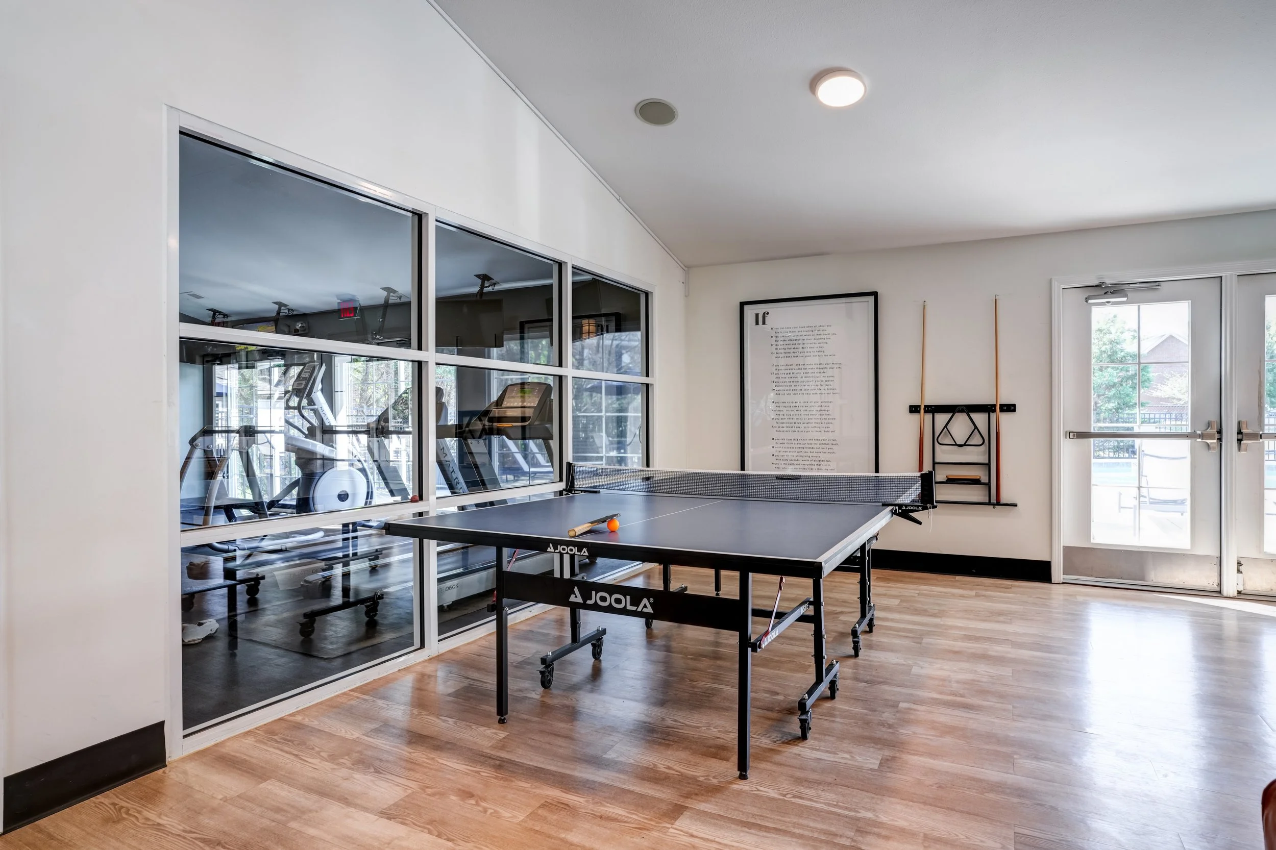 An indoor recreation room with a ping pong table in the center, a glass wall separating it from a workout area with treadmills, and a framed quote on the wall near the door.