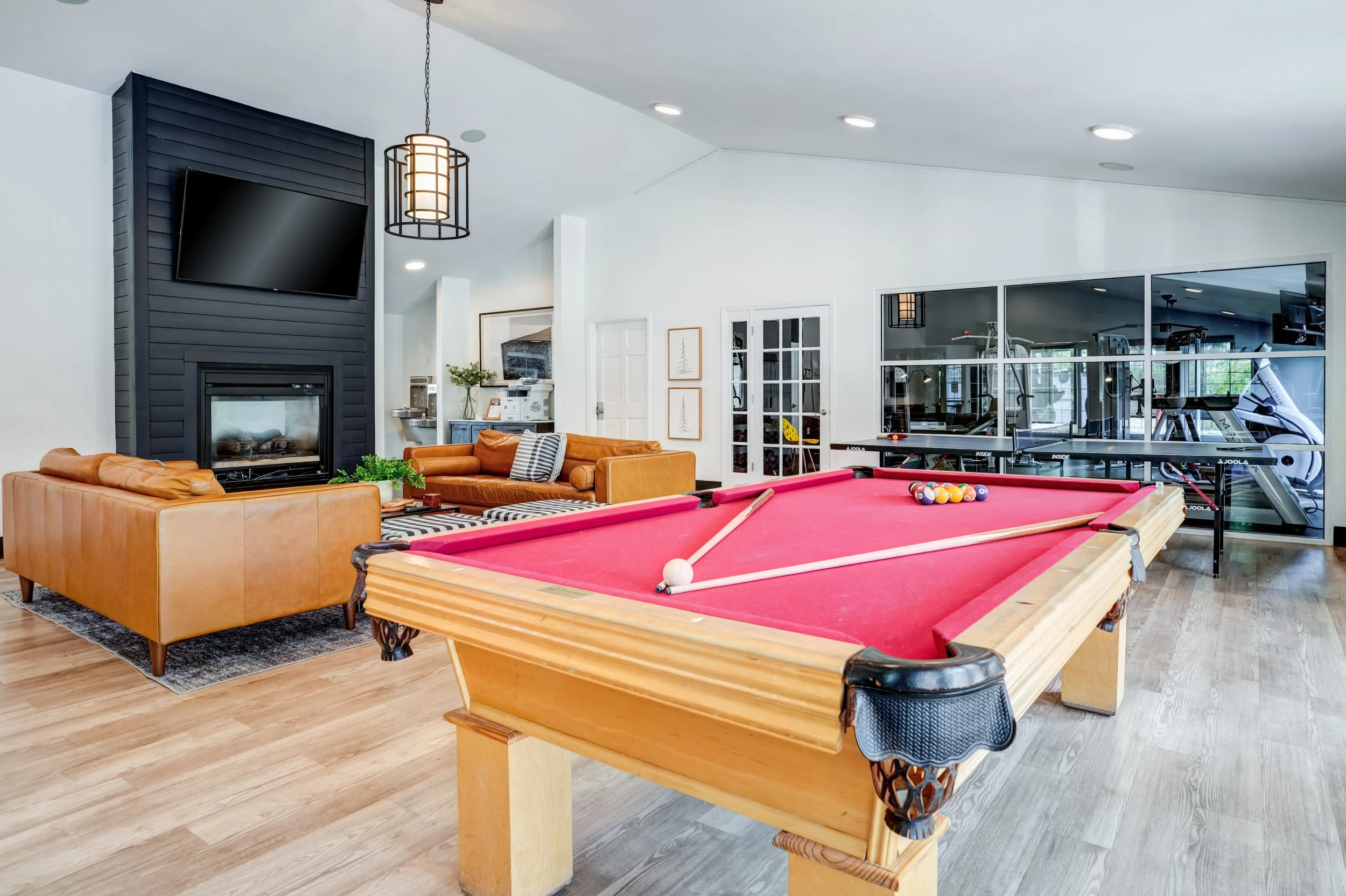 A recreation room with a red pool table, leather sofas, a large television mounted above a black fireplace, and a gym area with exercise equipment separated by glass windows.