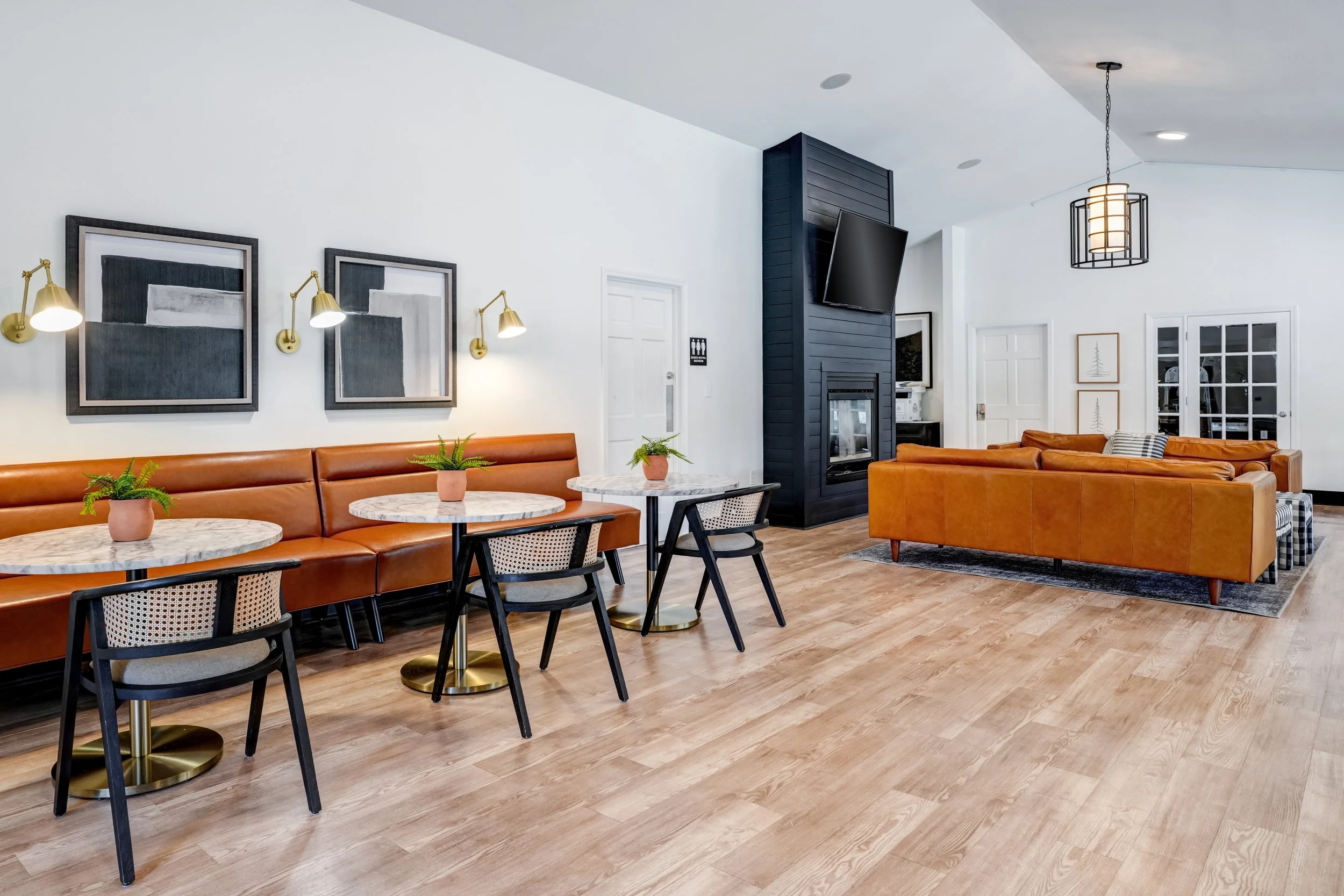 Modern clubhouse with white walls, brown leather couches, black-framed artwork, marble-topped tables with small green plants, and a black wood-paneled fireplace with mounted TV, illuminated by a hanging lantern and built-in ceiling lights.