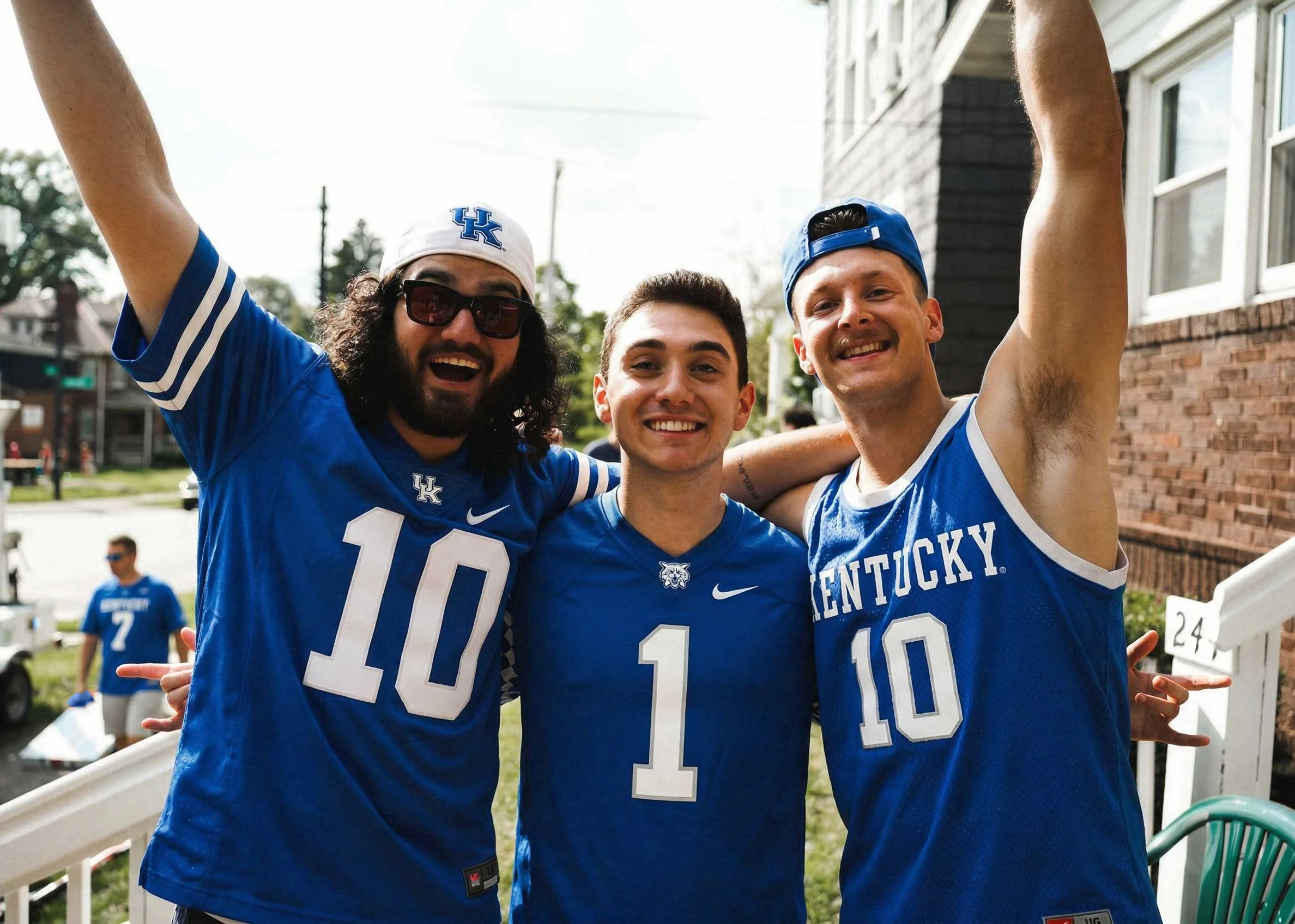 Three smiling young men dressed in Kentucky Wildcats sports jerseys and caps, celebrating outdoors on a sunny day with friends in the background.