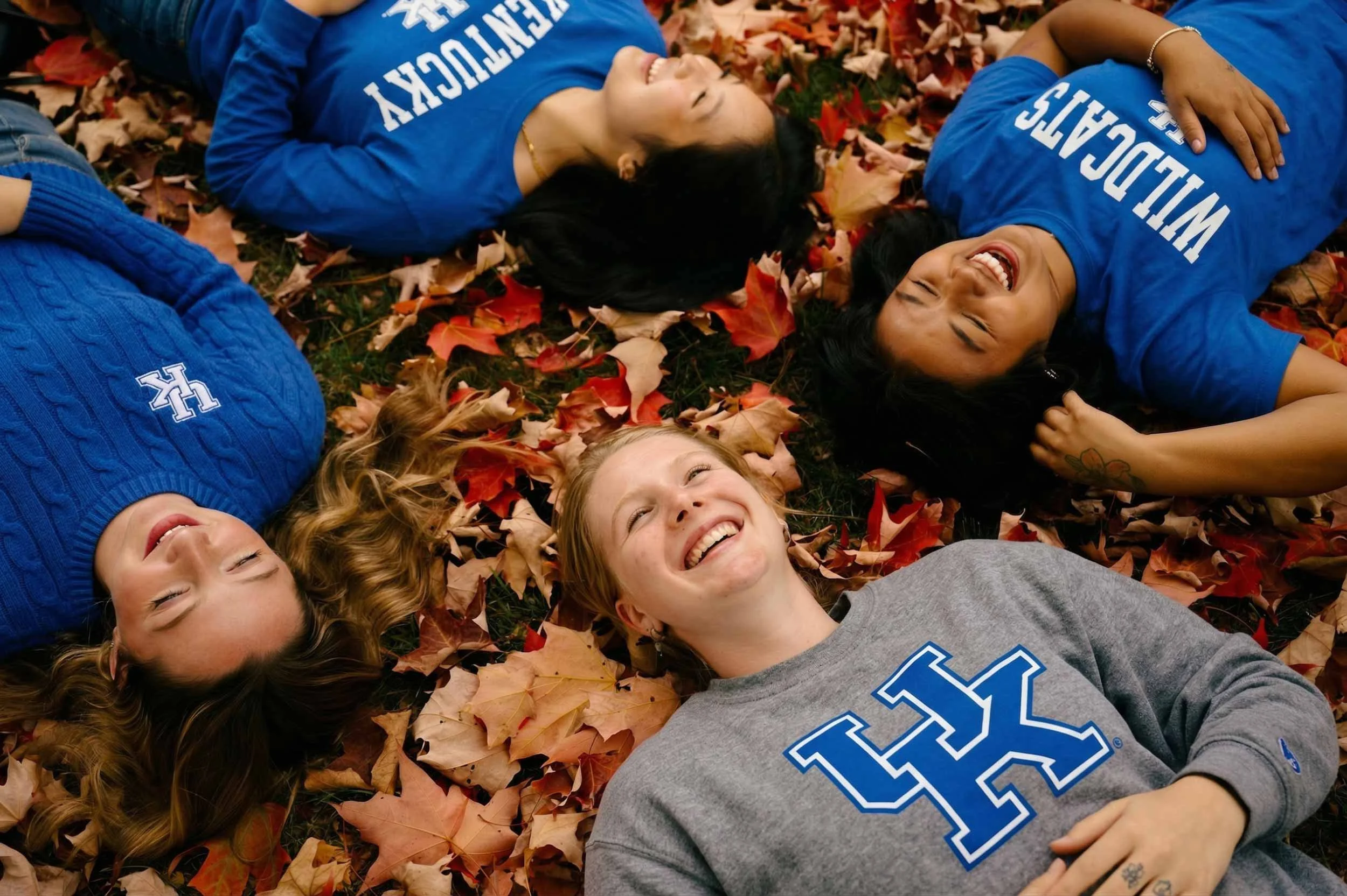 A group of people lying on a bed of fallen autumn leaves, smiling and laughing. They are wearing blue and gray shirts, some with writing on them, and are enjoying the outdoor fall scene.