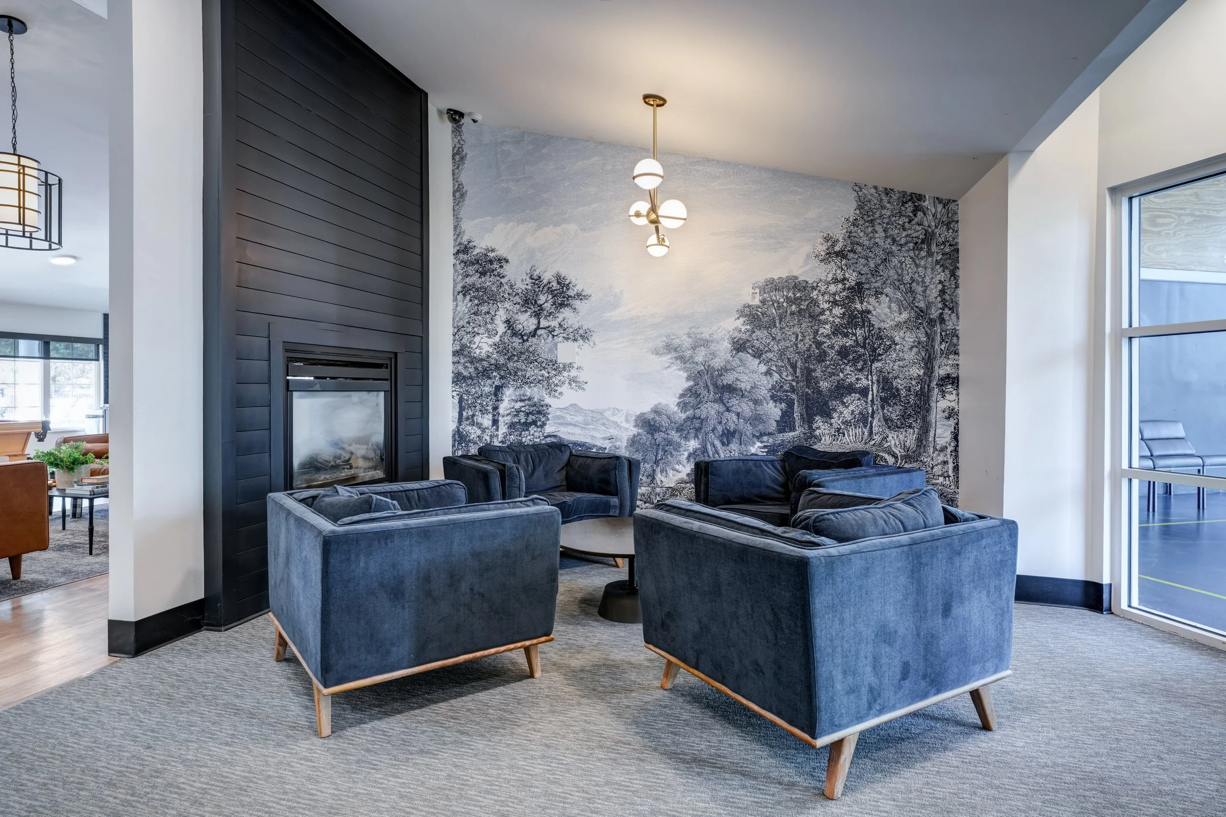 Modern living room featuring blue velvet armchairs around a round coffee table, with a large mural of trees and landscape on the wall, and a contemporary chandelier overhead.