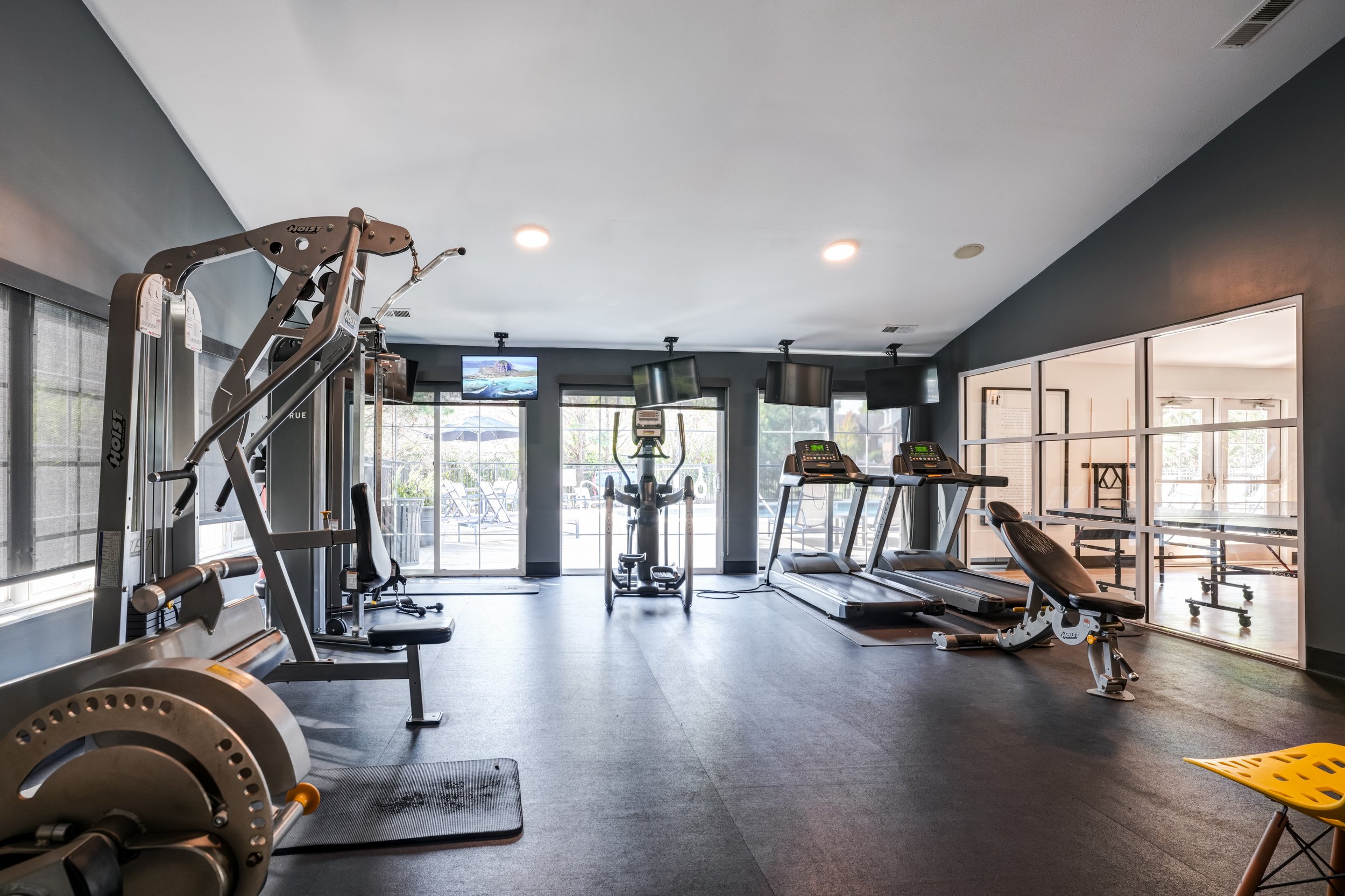 Interior of a modern gym with various workout equipment including treadmills, ellipticals, and weight machines, large windows, and mounted TVs.