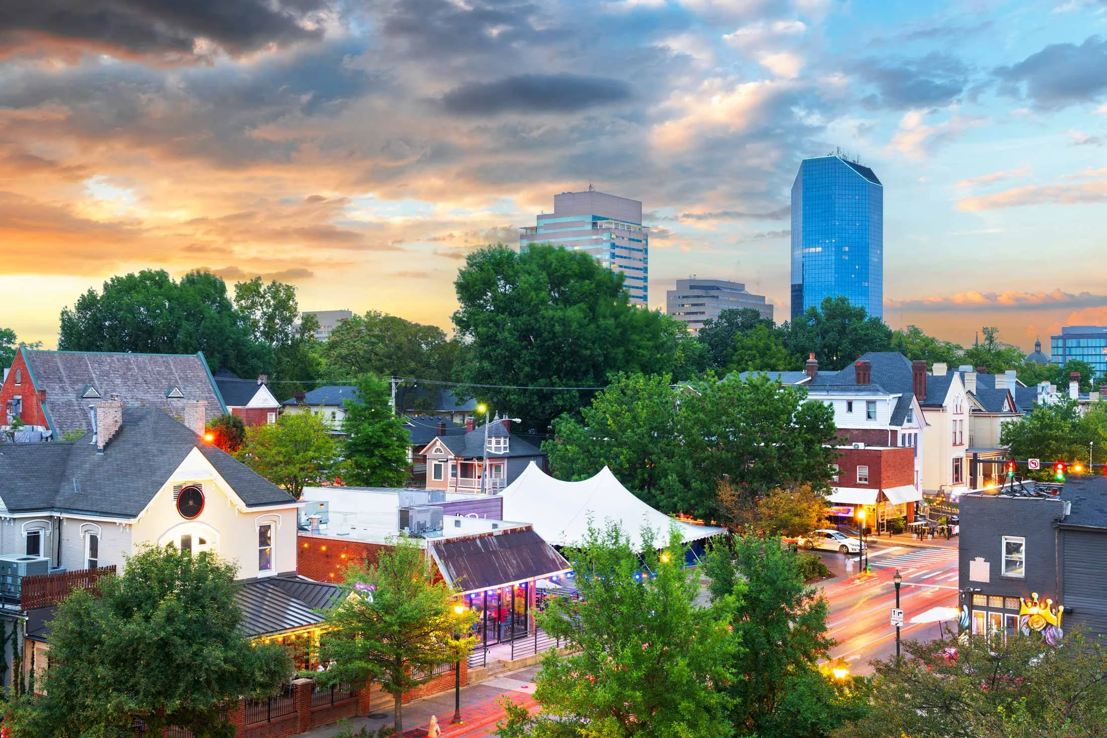 Cityscape during sunset with tall modern glass buildings, residential houses, lush green trees, and a busy street with moving vehicles.