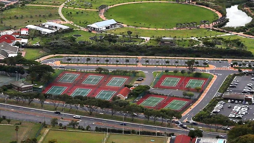 Multiple tennis courts with red and green surfaces, surrounded by parking lots, trees, and pathways in a park-like setting.