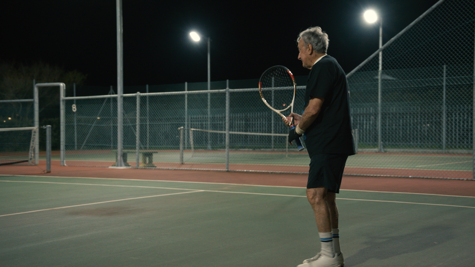An elderly man standing on a tennis court at night, holding a tennis racket, with floodlights illuminating the court and a chain-link fence in the background.