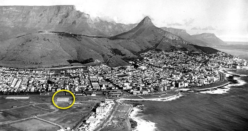 A black and white aerial photograph of a coastal city with a mountain range in the background. There is a stadium with a running track and a filled seating area, and an area outlined in yellow nearby.