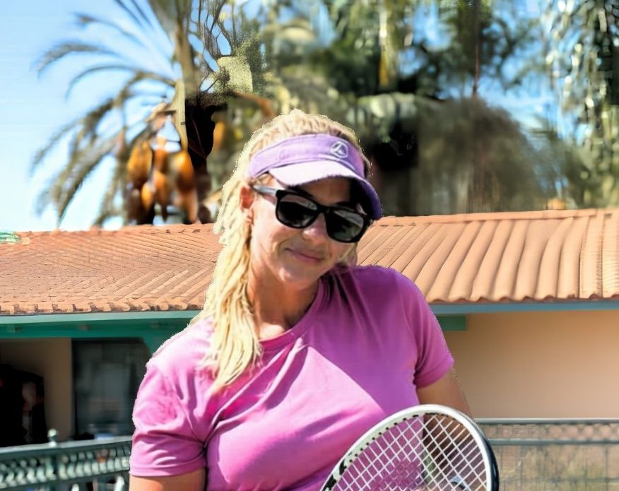 A woman wearing sunglasses, a purple cap, and a purple shirt holding a tennis racket outdoors with palm trees and a building with a tiled roof in the background.