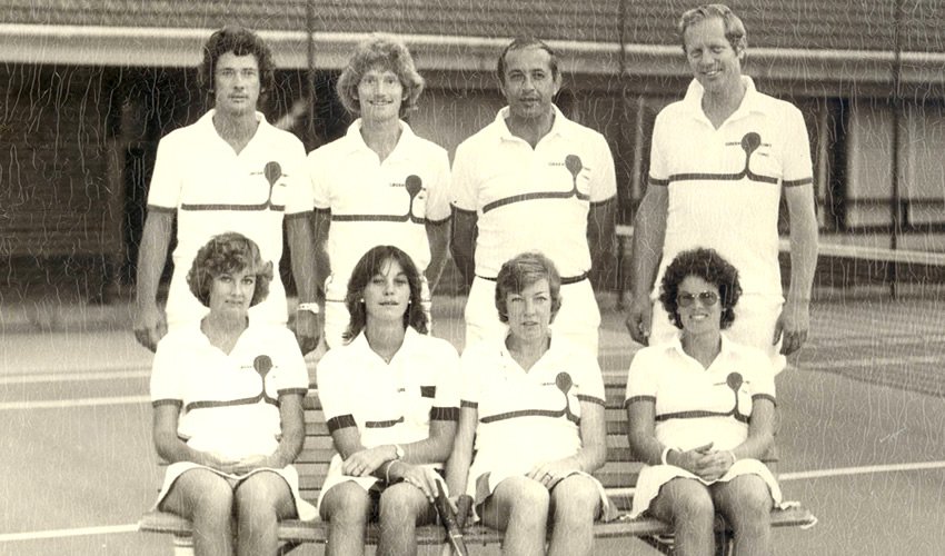 Group of seven tennis players, five women seated and two men standing, on a tennis court, all wearing tennis uniforms with striped shirts.