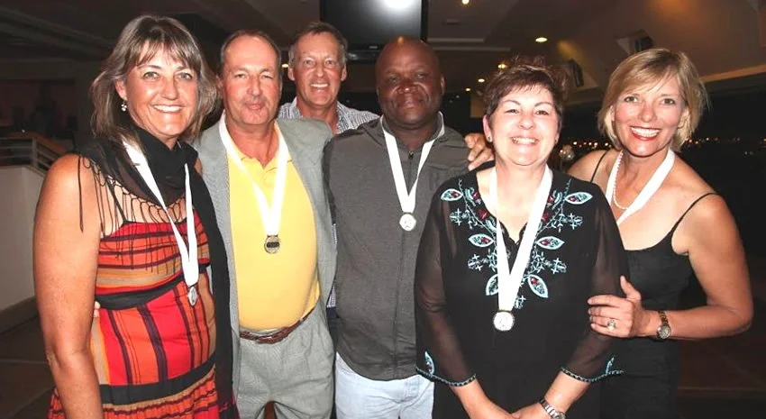 Six people at an indoor event, all wearing medals around their necks, smiling for the photo.