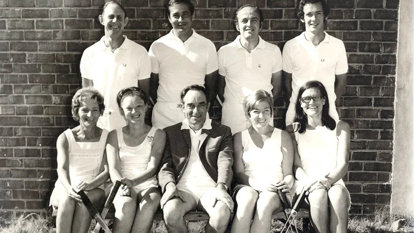 Group of nine people, five women and four men, posing outdoors against a brick wall, all smiling. The women are seated in the front row, and the men are standing in the back row.