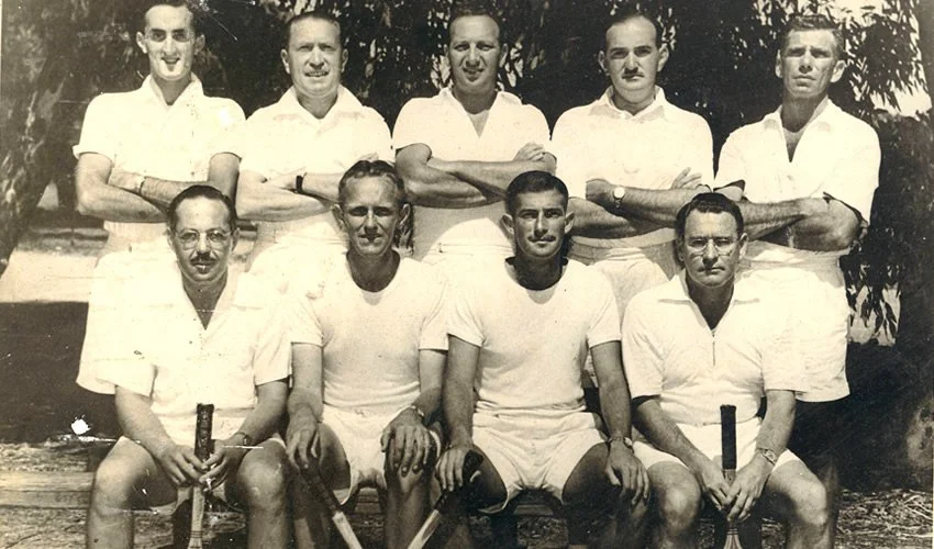 Black and white photograph of nine young men in light-colored sports clothing, posing outdoors with some holding hockey sticks.