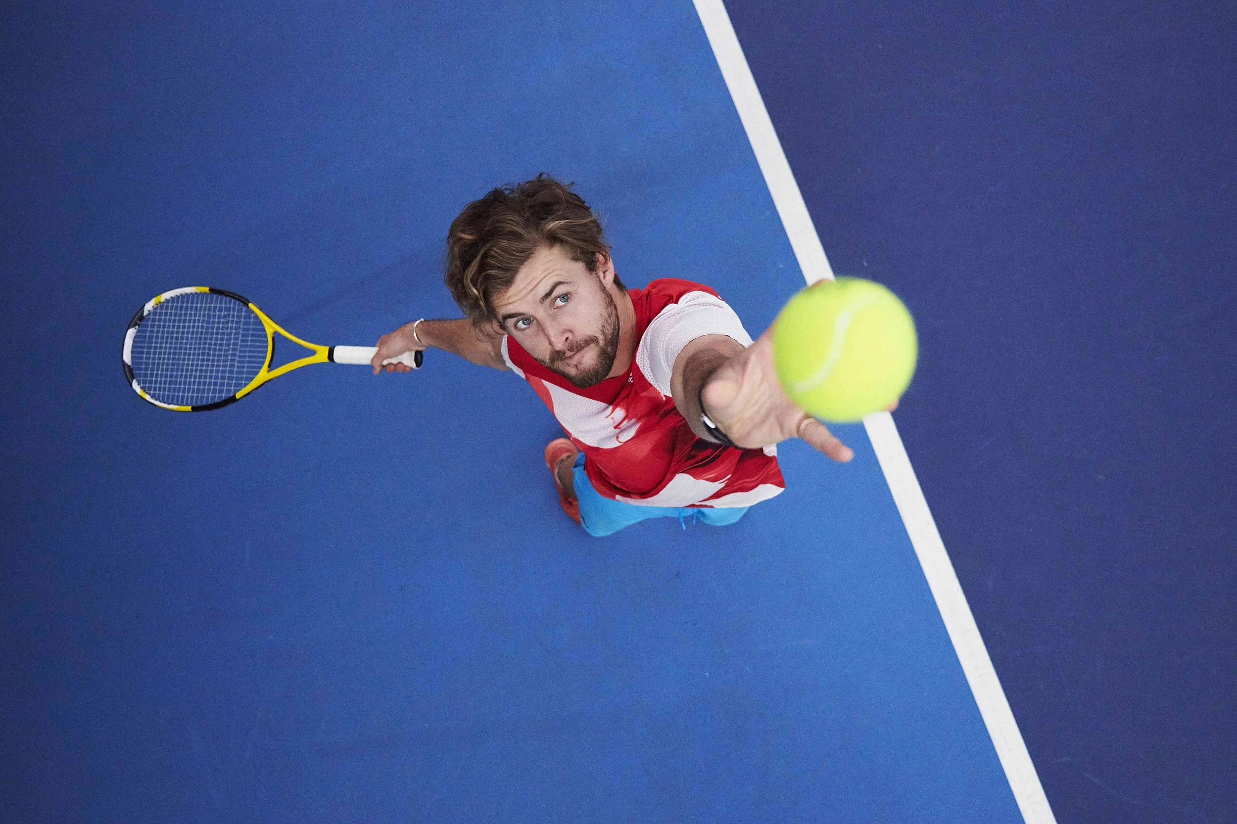 Above view of a man with brown hair and beard playing tennis on a blue court, holding a yellow tennis racket in his left hand and reaching out with his right hand to hit a yellow tennis ball.