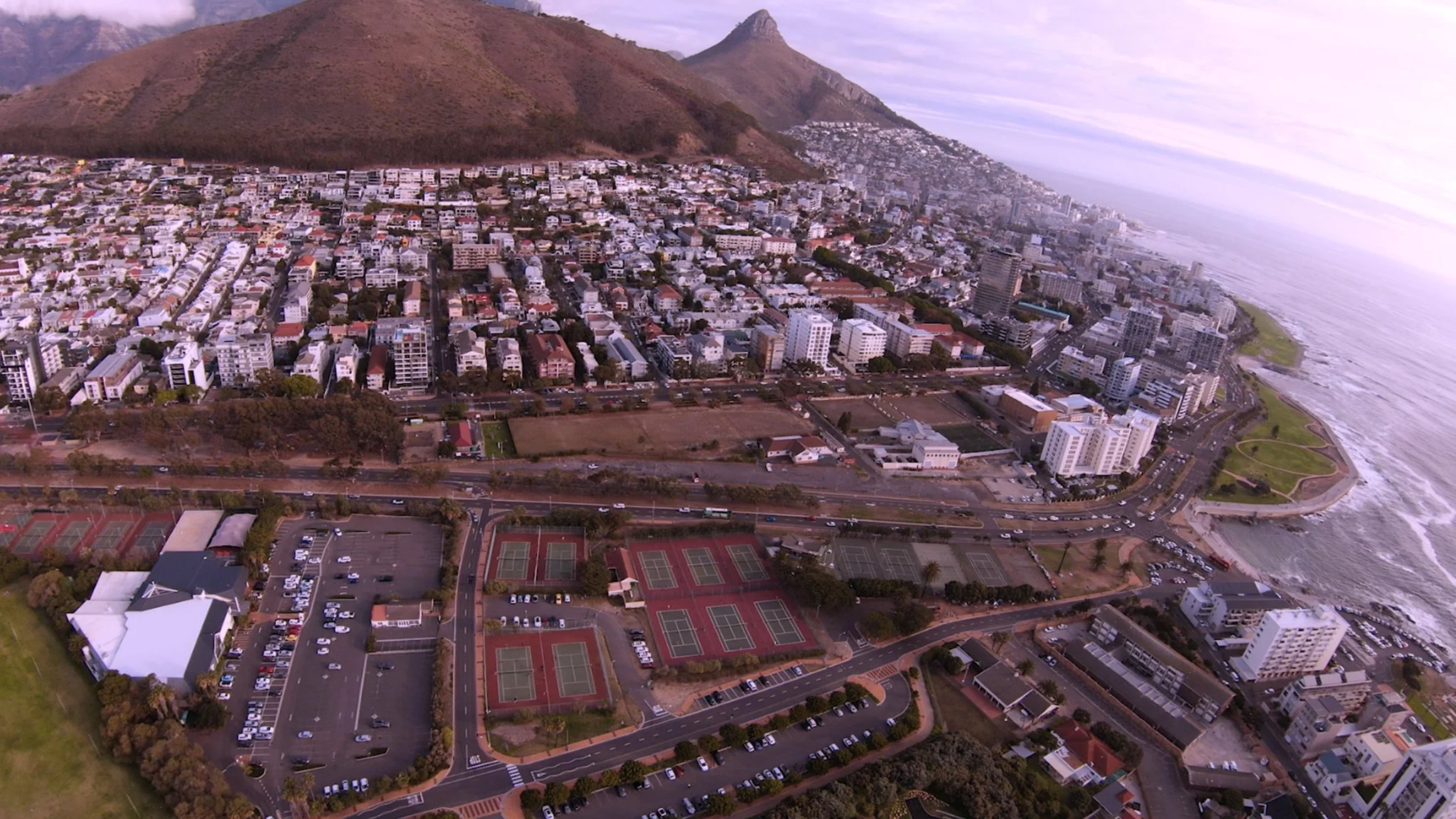Aerial view of a coastal city with a mix of high-rise buildings, residential neighborhoods, and tennis courts adjacent to the ocean, with mountains in the background.