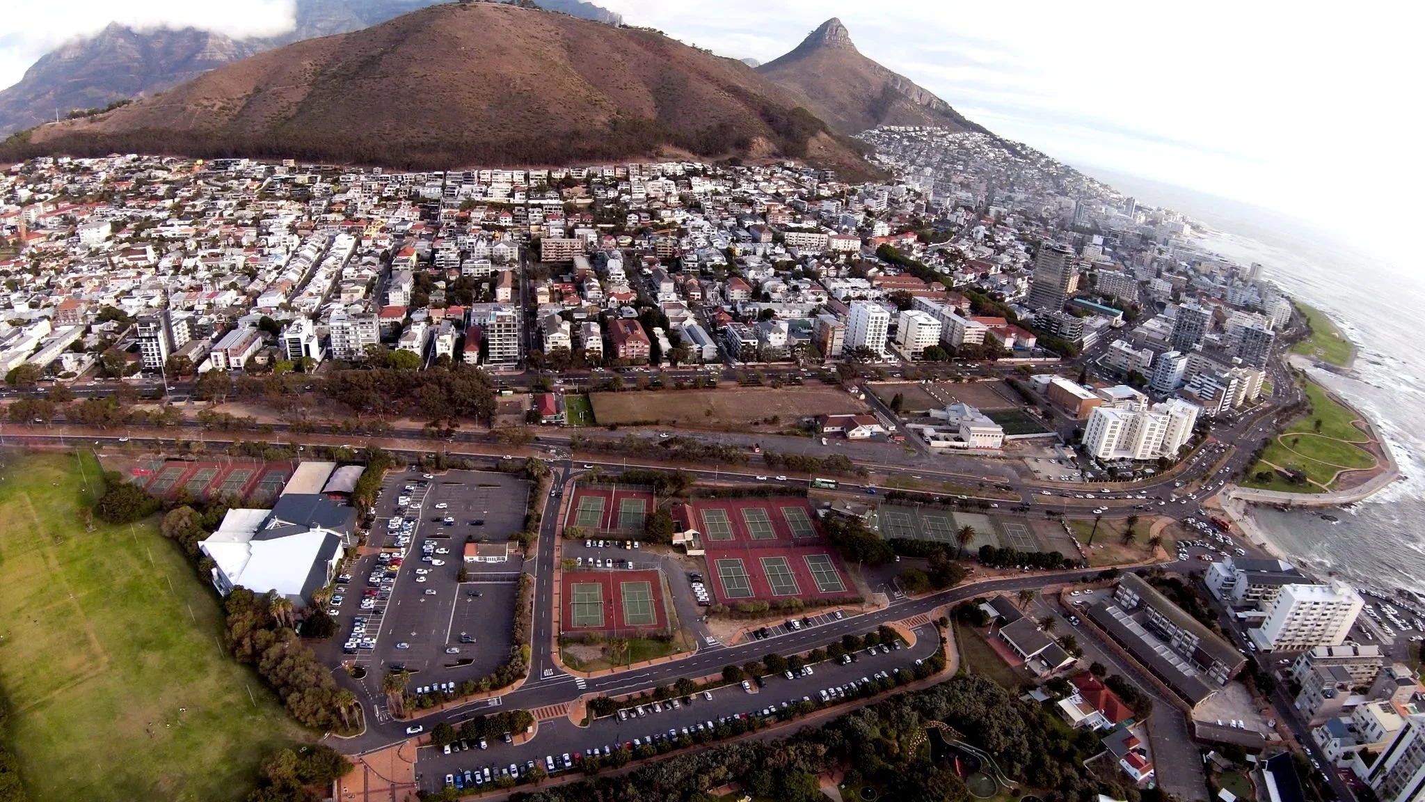 Aerial view of a coastal city with densely packed buildings, a soccer field, tennis courts, and a waterfront with a promenade.