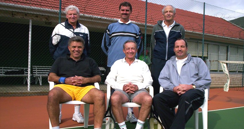 Six men on a tennis court, three sitting in chairs and three standing behind them, with a net and building in the background.