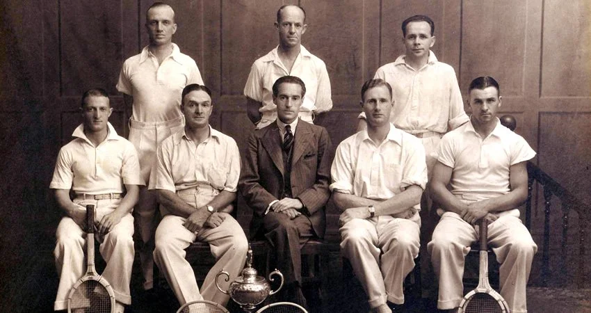 Black and white photo of eight men, four standing in the back and four seated in the front, with a wooden-paneled wall behind them. The seated men are holding tennis rackets and there is a trophy in front of them.