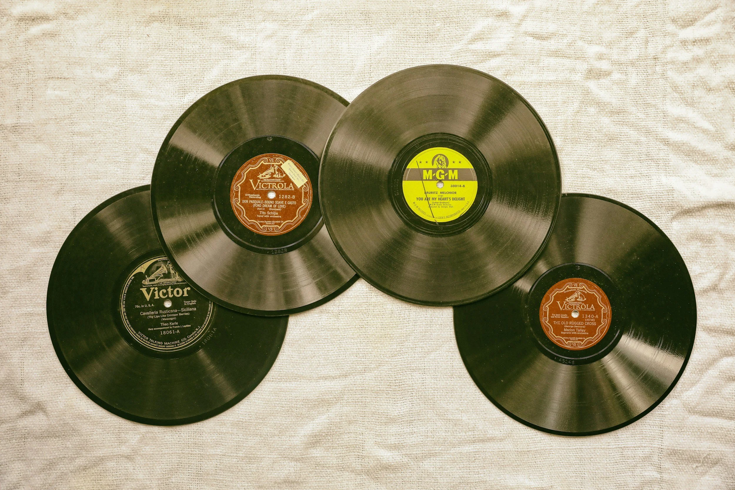 Collection of vintage black vinyl records with colorful labels, placed on a white textured fabric surface.