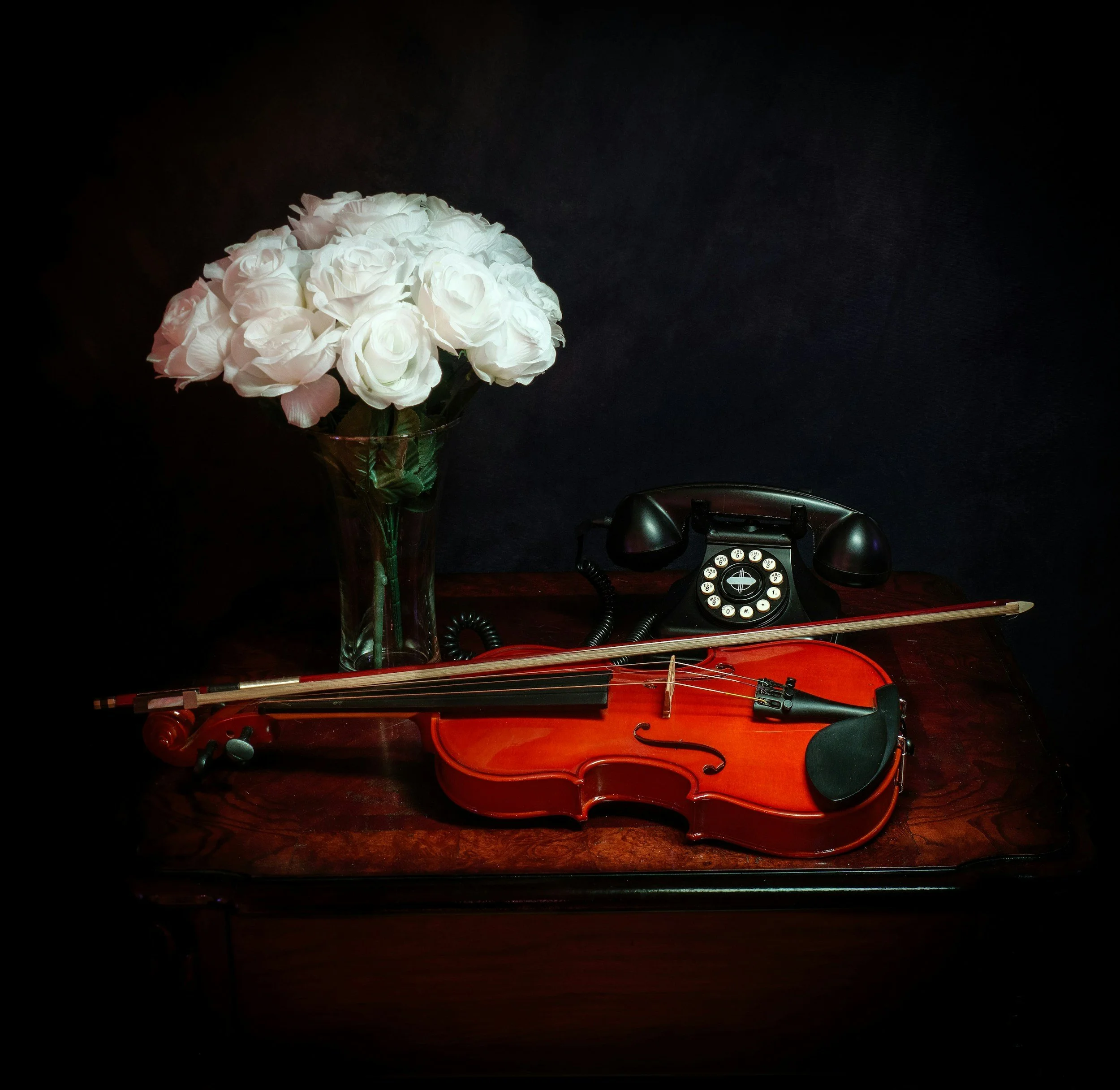 A bouquet of white roses in a glass vase on a dark surface, with a violin and a black phone nearby.