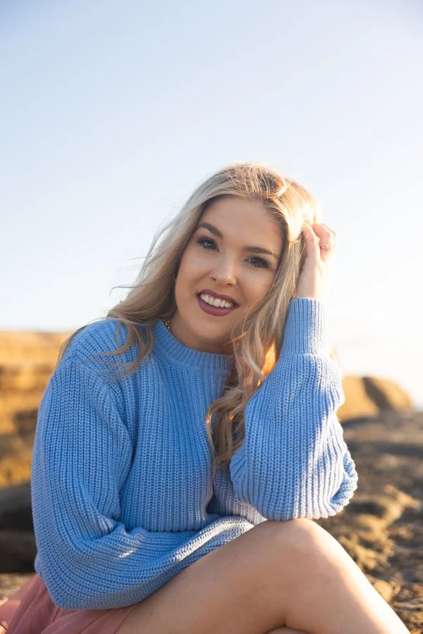 A young woman with blonde hair wearing a blue sweater, sitting outdoors on rocks, smiling at the camera with a clear sky background.