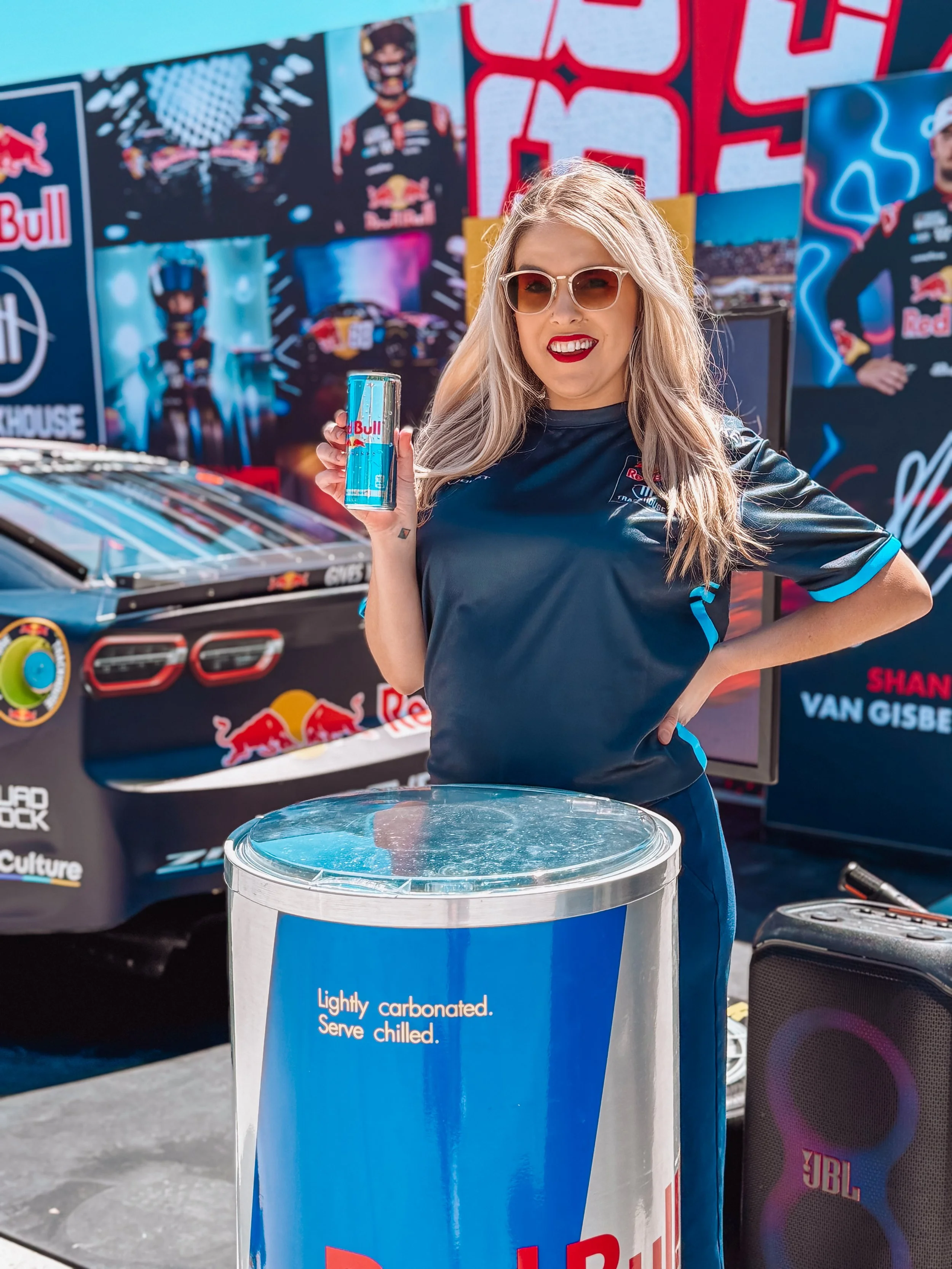 A woman with blonde hair, sunglasses, and red lipstick is smiling and holding a Red Bull energy drink can. She is dressed in a dark blue Red Bull racing shirt. Behind her, there is a race car with Red Bull branding and posters featuring race drivers and Red Bull logos. She is standing next to a blue trash bin with text that reads, 'Lightly carbonated. Serve chilled.'