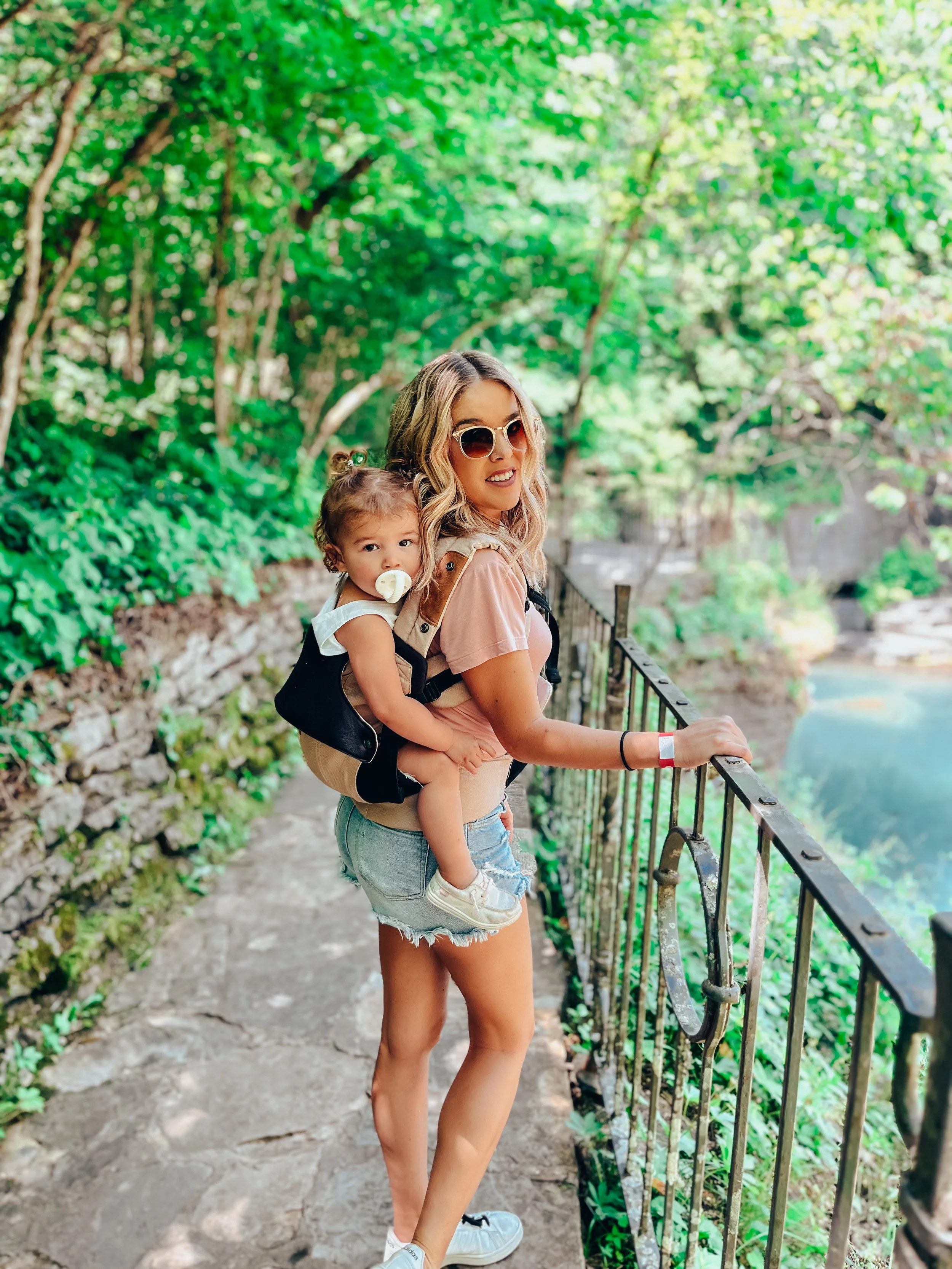 A woman carrying a young girl on her back while standing on a nature trail with green foliage in the background. The woman is wearing sunglasses, a pink t-shirt, and denim shorts. The girl has a pacifier in her mouth and is dressed in a white top and shorts. They are next to a metal railing by a body of water.